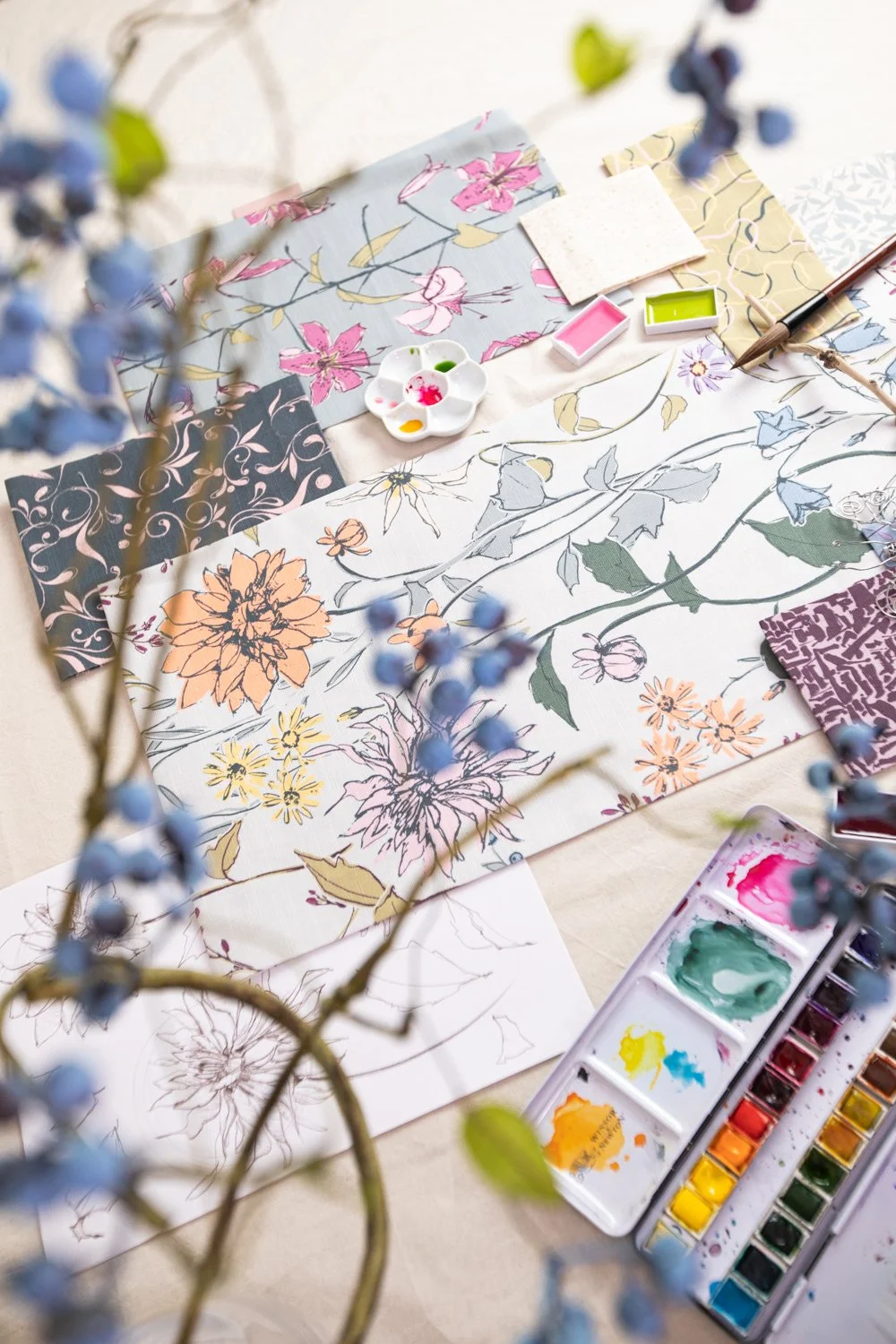 Artistic watercolor painted floral fabric samples and watercolor paints on a table, with some purple flowers in the foreground.