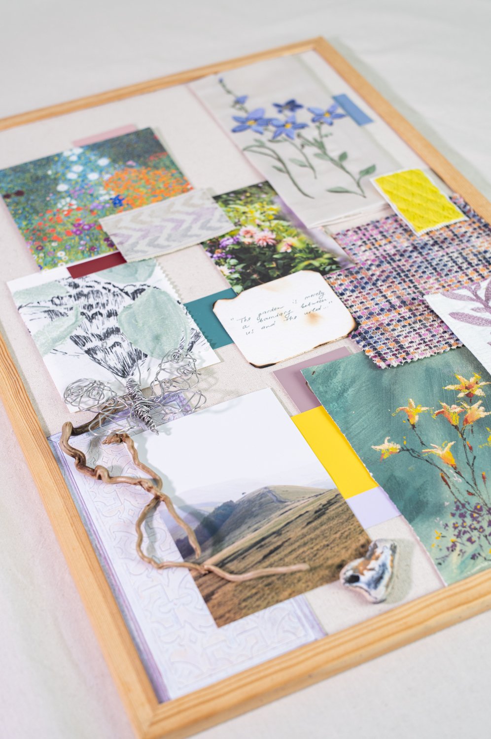 A mixed-media collage in a wooden frame containing photos, fabric swatches, small notes, drawings of flowers, a wire butterfly, and a piece of twisted wood, arranged on a light-colored surface.
