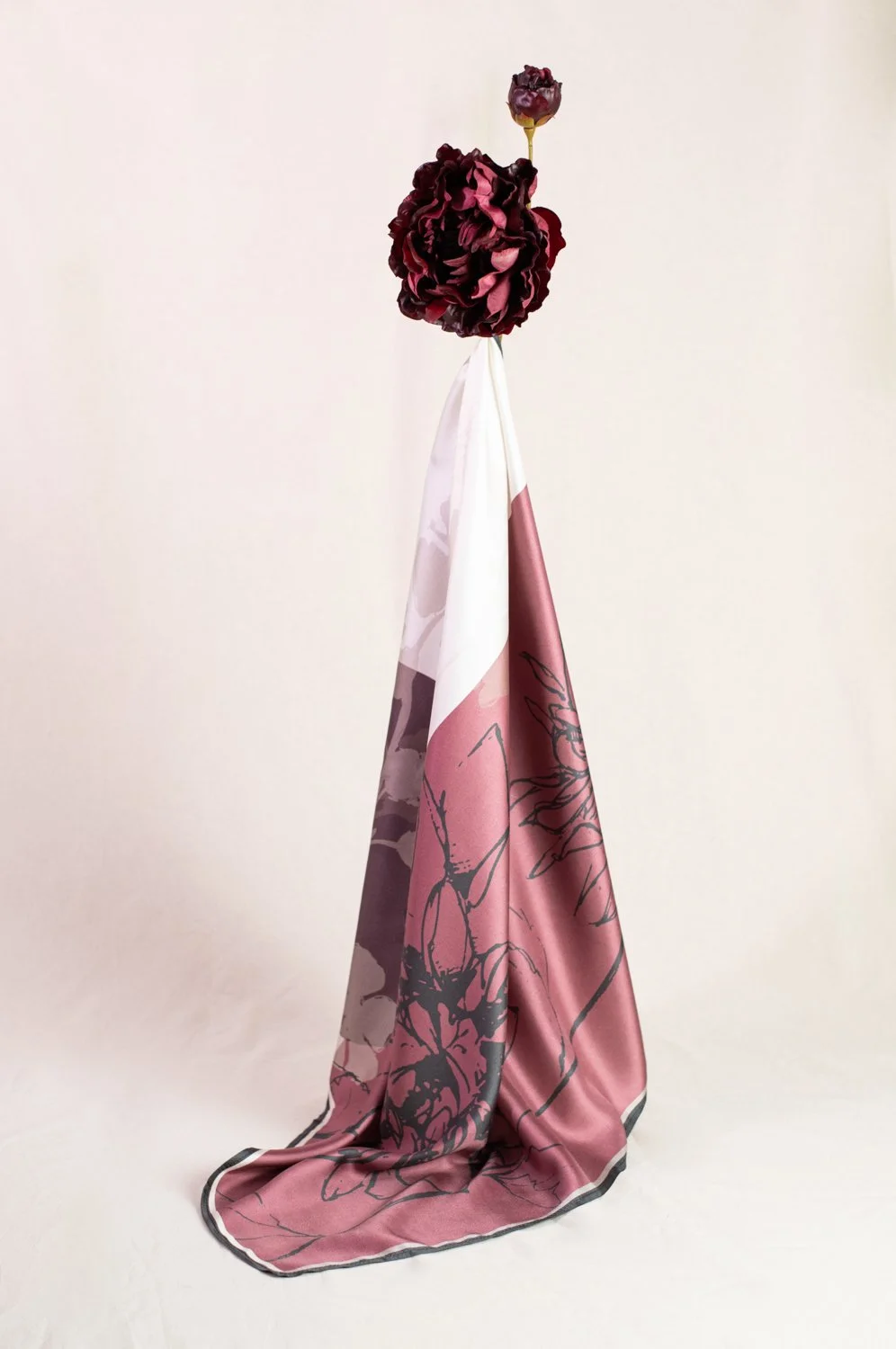 A dark red flower is placed on top of a scarf with floral print draped over a stand, against a plain light-colored background.