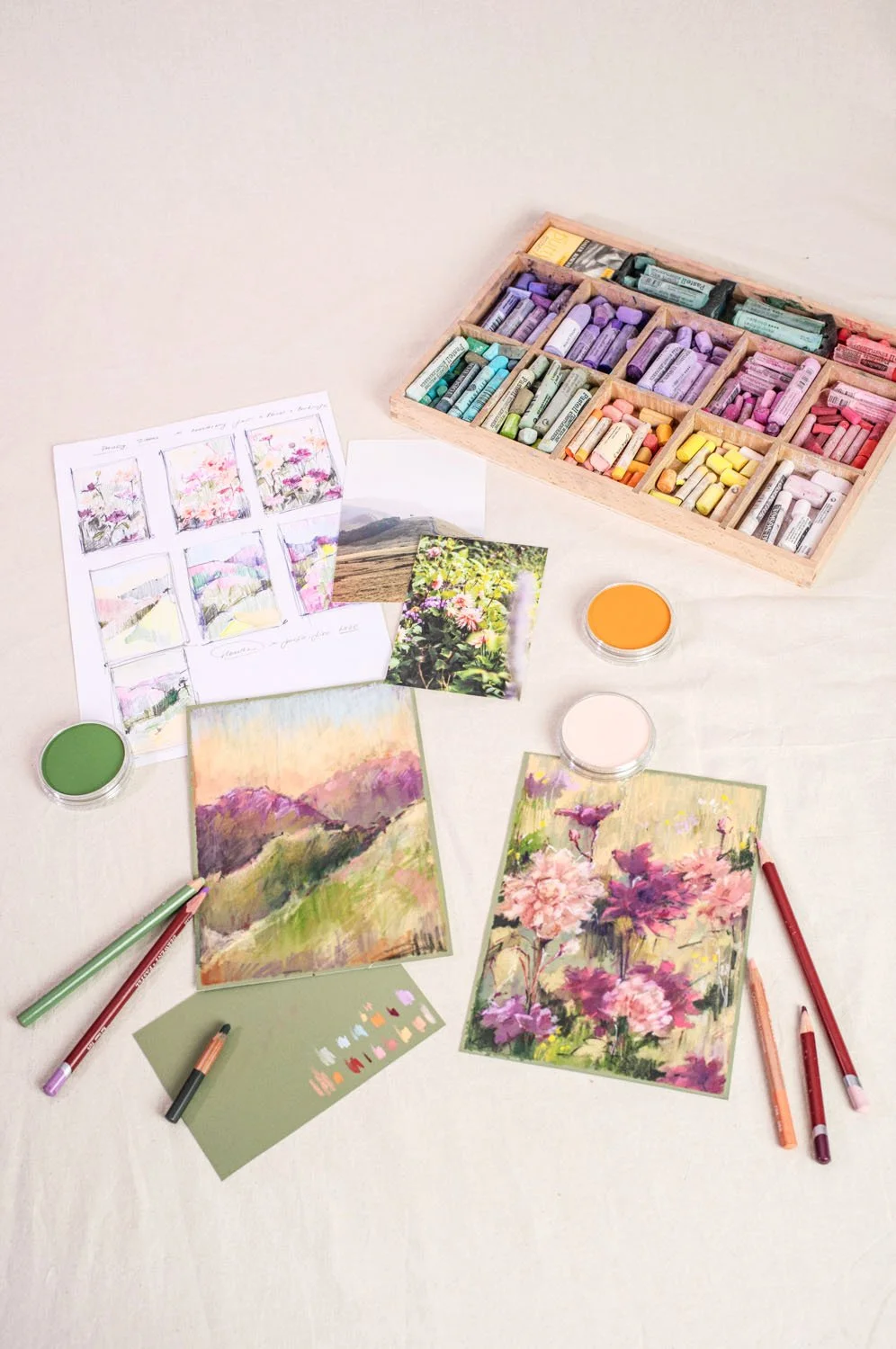 Art supplies including pastel chalks in a wooden box, watercolor paintings, printed photos, paint containers, markers, and color swatches on a white surface.