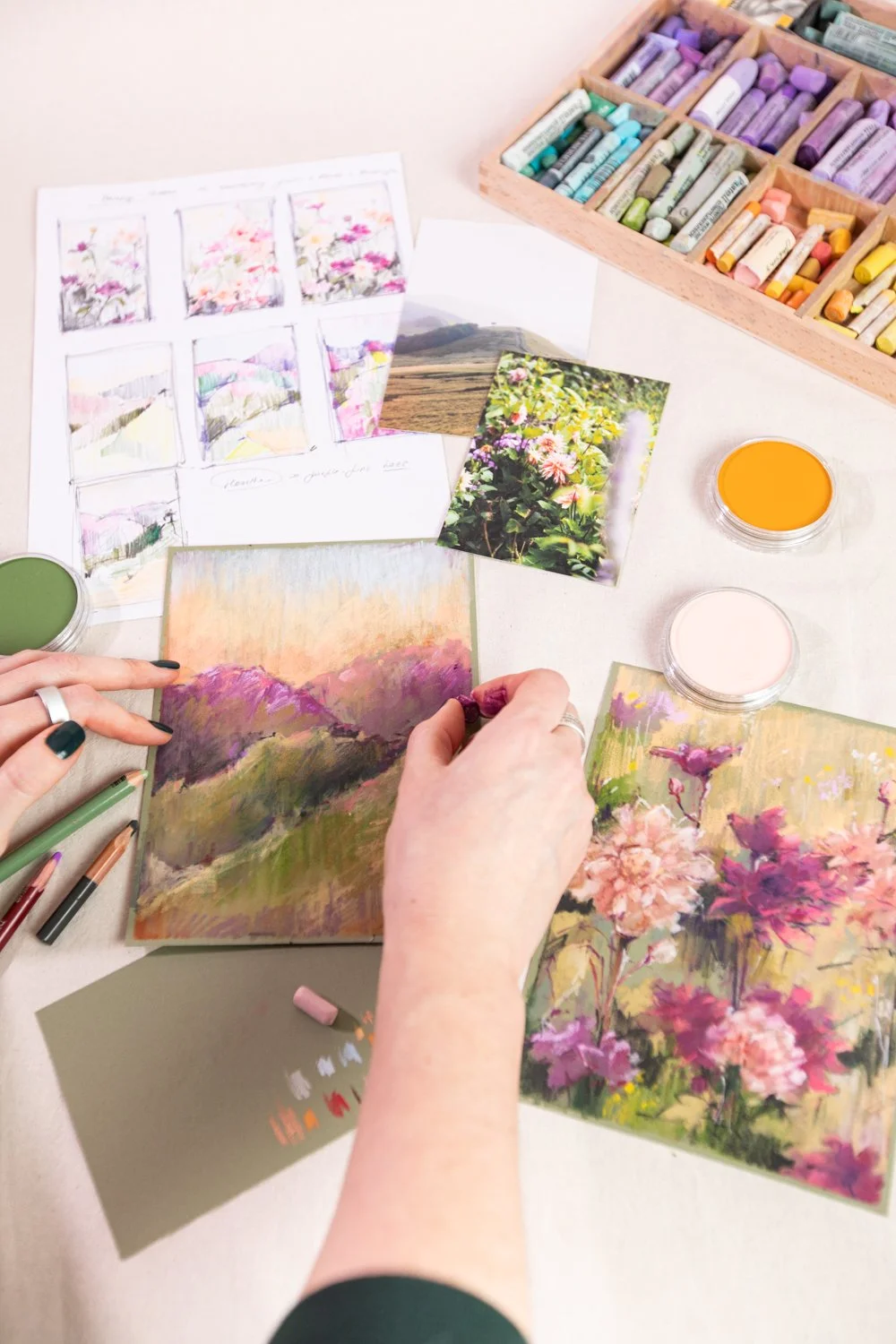 An artist creating landscape paintings with pastel colors. Various reference photos of flowers and nature scenes are on the table. Pastel chalks, paints, and art supplies are visible, along with finished or in-progress landscape artworks featuring mountains, flowers, and fields.
