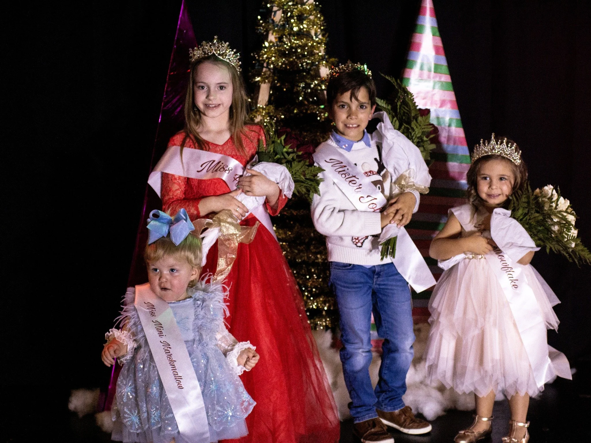 Four children dressed in fancy clothes and tiaras, holding bouquets of flowers, standing in front of a decorated Christmas tree with a black background.