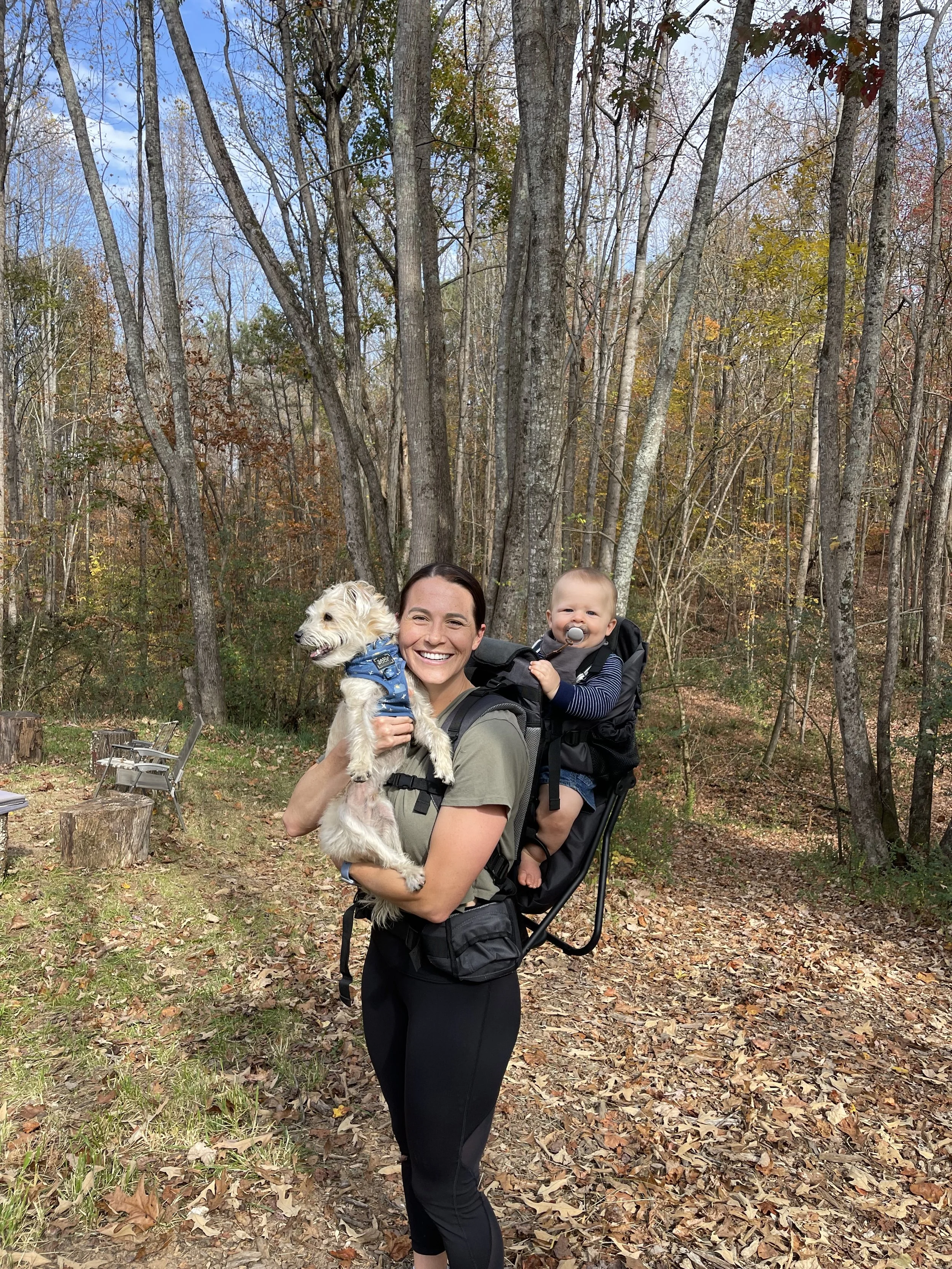 A smiling woman carrying a small dog and a baby in a backpack carrier in a wooded park during fall.