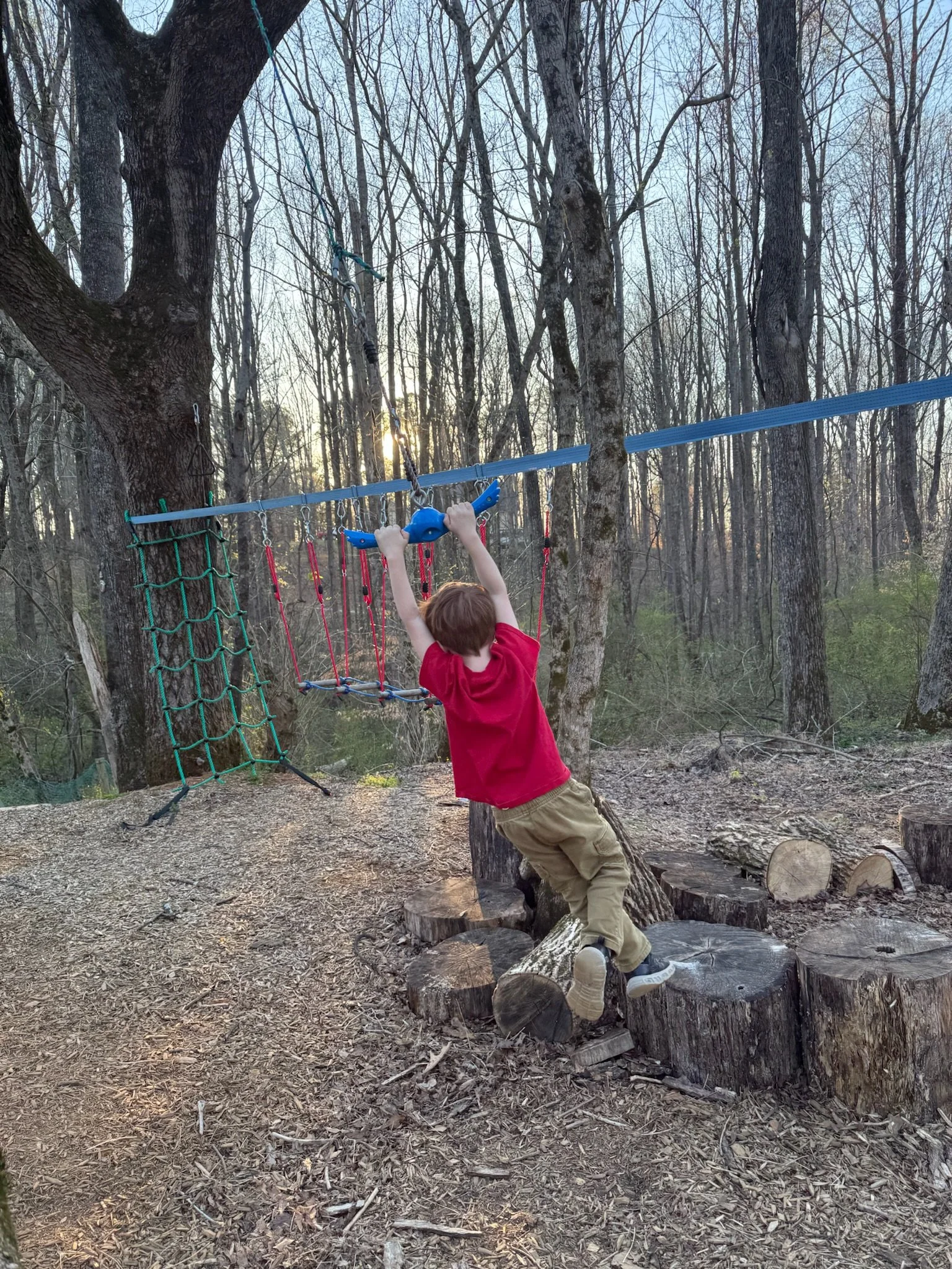 Child wearing a red shirt and tan pants playing on a wooden balance beam in a wooded outdoor area during sunset.