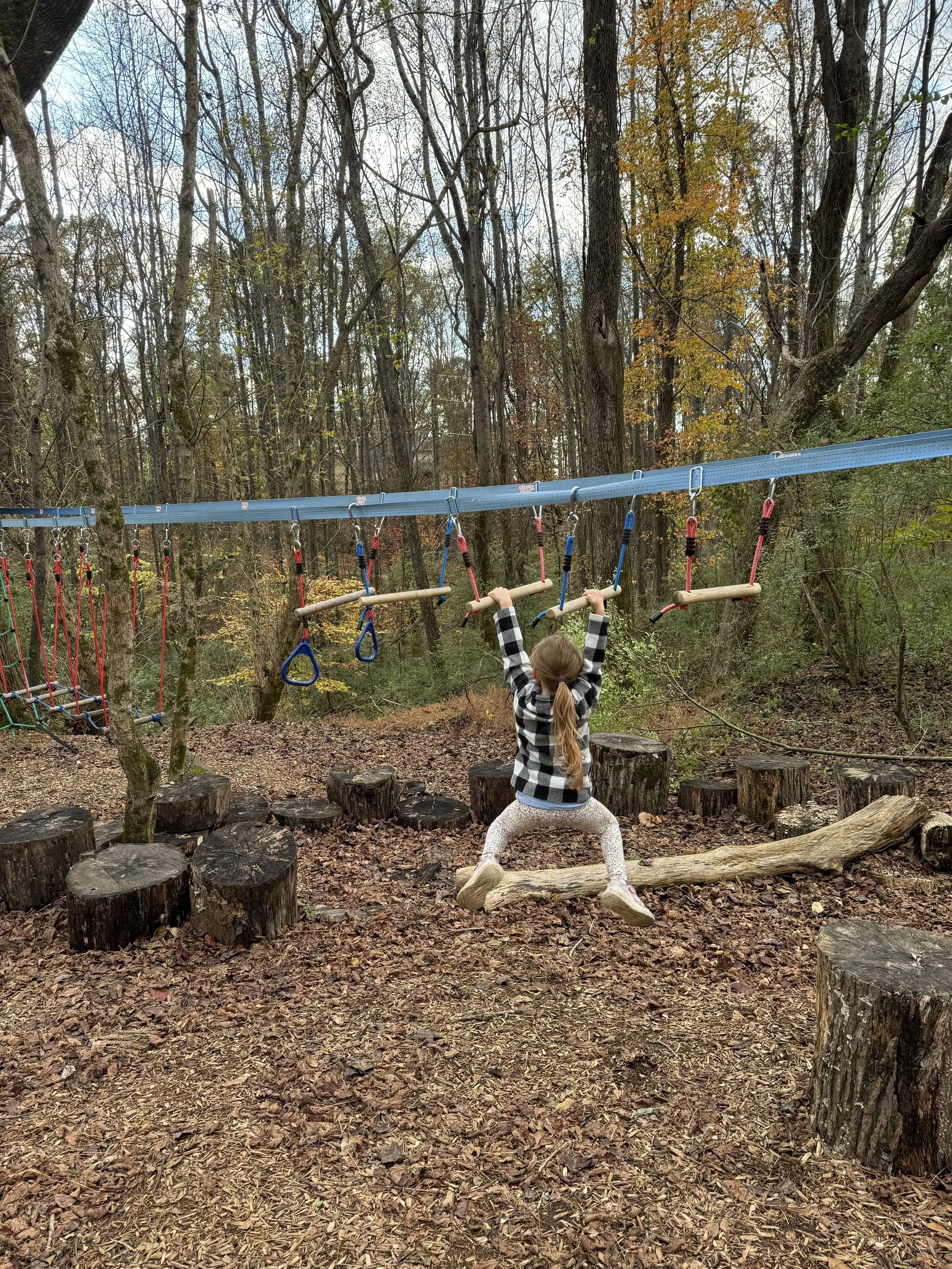 A child with a ponytail, wearing a black and white plaid jacket and white leggings, swings on a log in a forested playground area during fall, with some trees having orange leaves and others bare.
