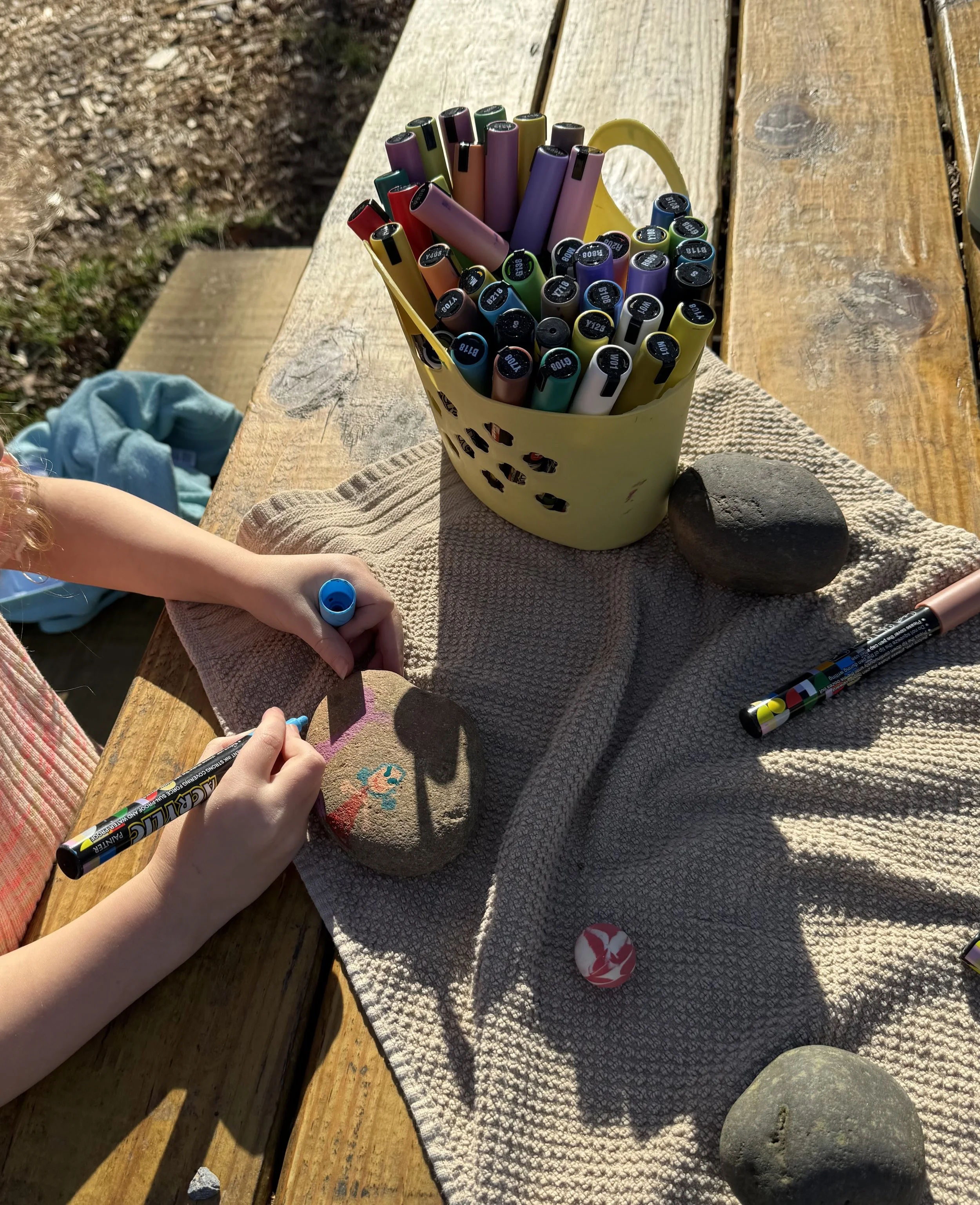 A person decorating a painted rock with markers on a wooden table outdoors, with a basket of colorful markers and two rocks nearby.