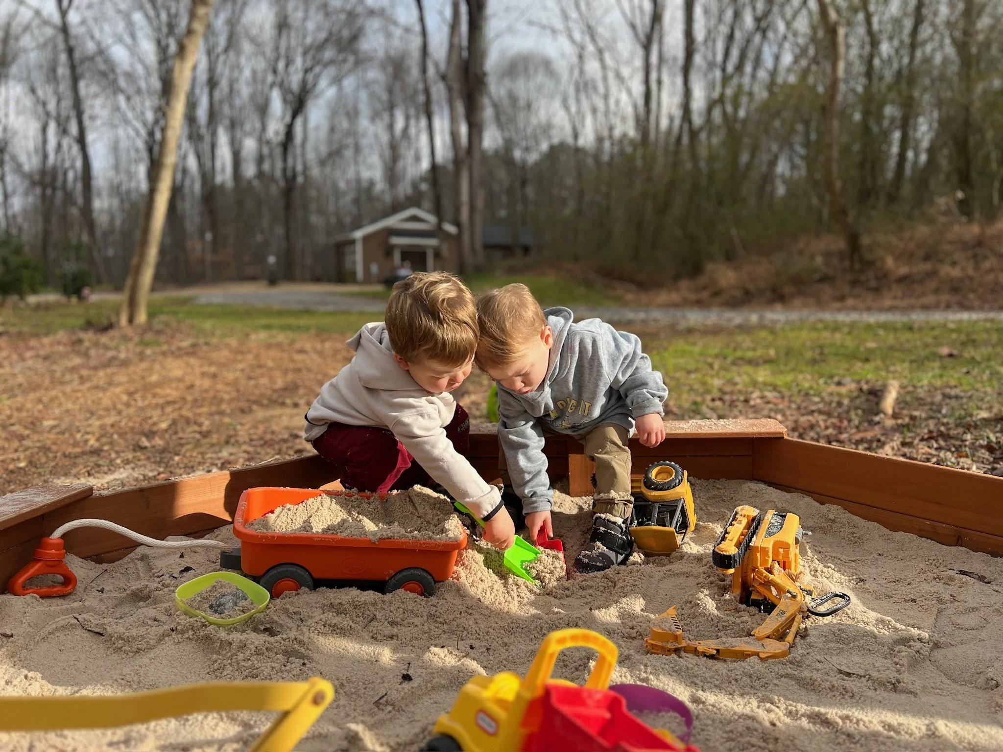 Two young children play in a sandbox outdoors with toy trucks and shovels, surrounded by trees and a small building in the background.