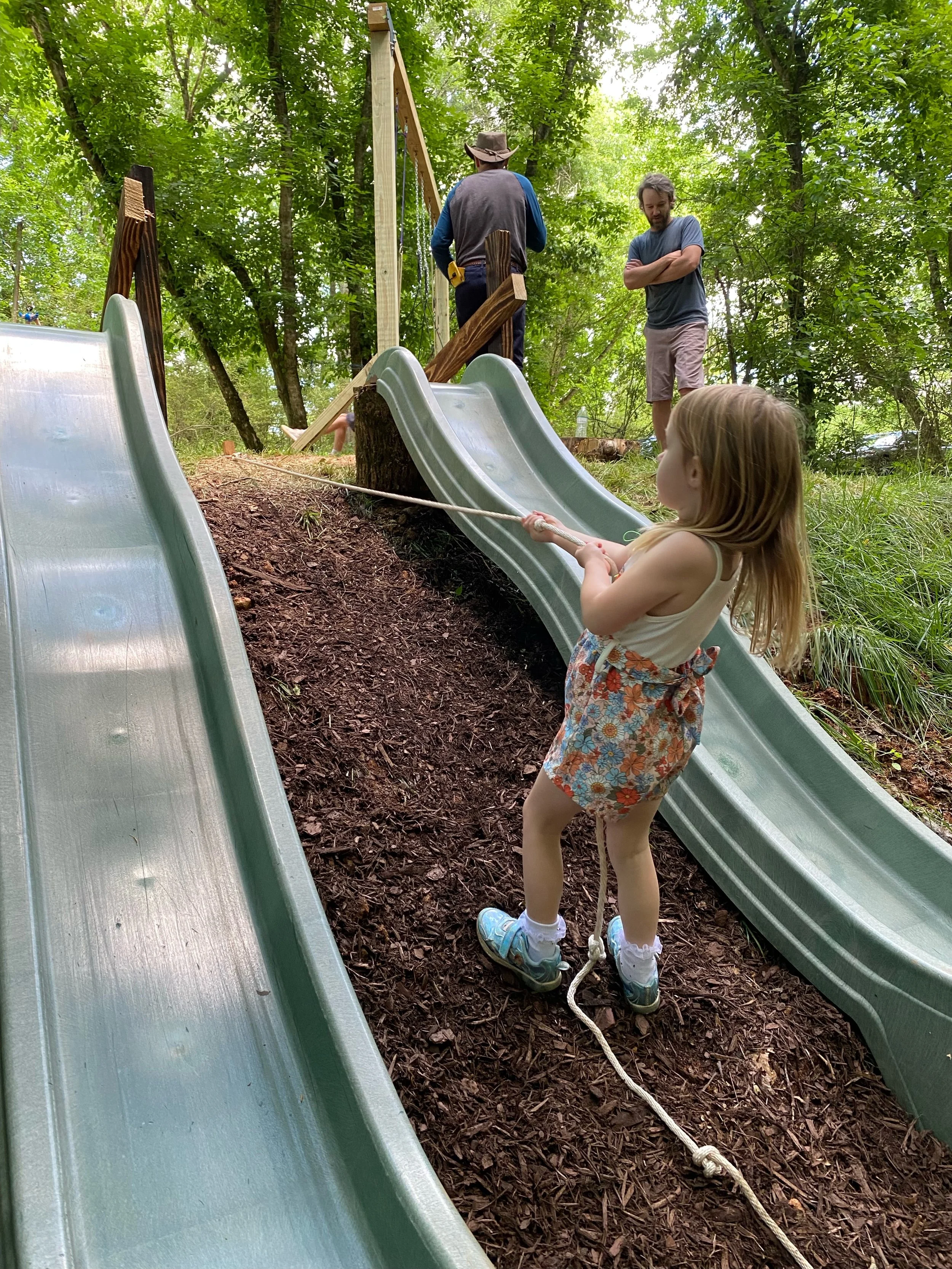 A young girl on a rope ladder on a wooden playground structure in a forest, with two men working on the treehouse above.