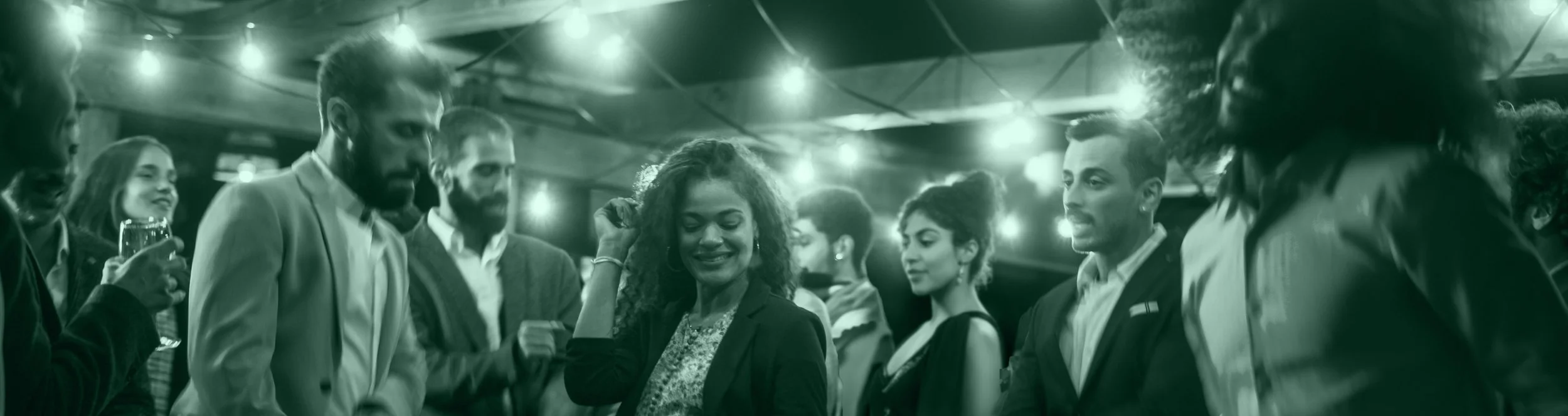 Group of diverse people at a social gathering, illuminated by string lights, engaging in conversation and enjoying drinks.