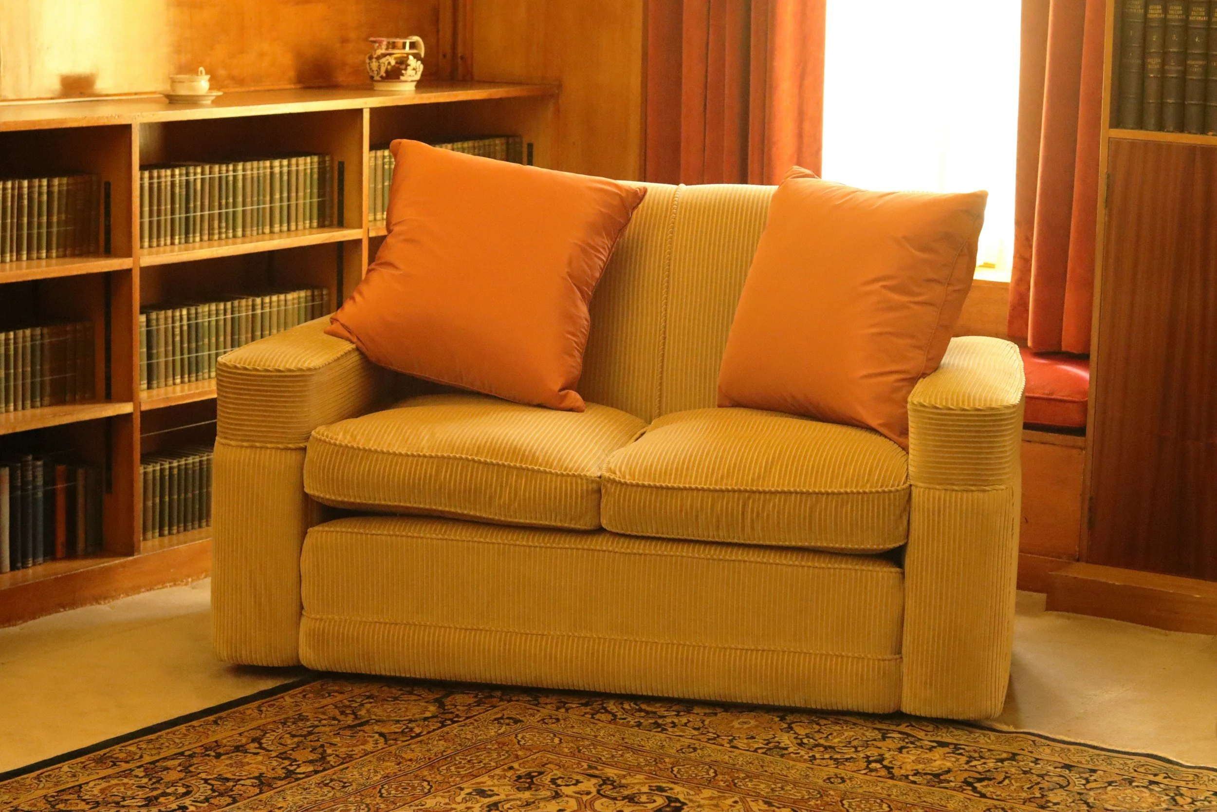 A yellow striped loveseat with two large rust-colored pillows. Behind the loveseat are wooden built-in shelves filled with books and some decorative items, with a window and curtains providing natural light.