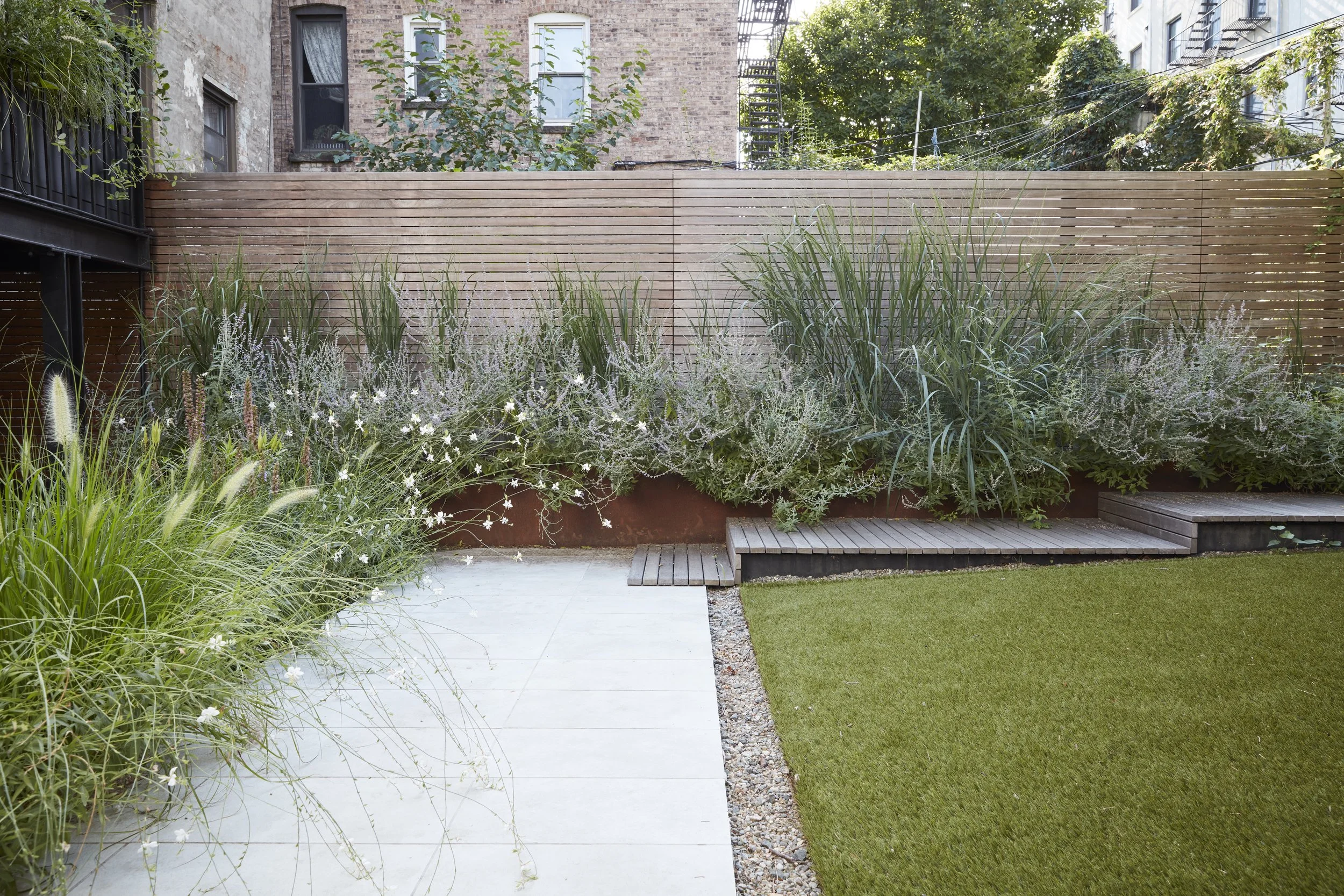 Urban backyard with a green lawn, white stone patio, and a raised garden bed filled with tall grasses and plants, enclosed by a wooden fence.