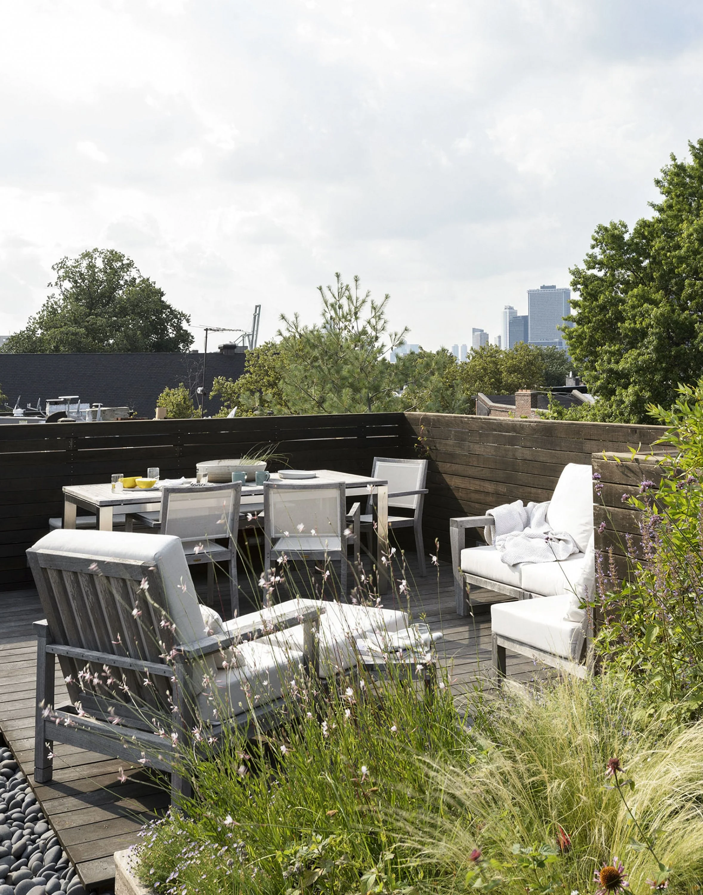 A rooftop patio with white outdoor furniture, including armchairs, a dining table, and chairs, surrounded by lush greenery and city skyline in the background.