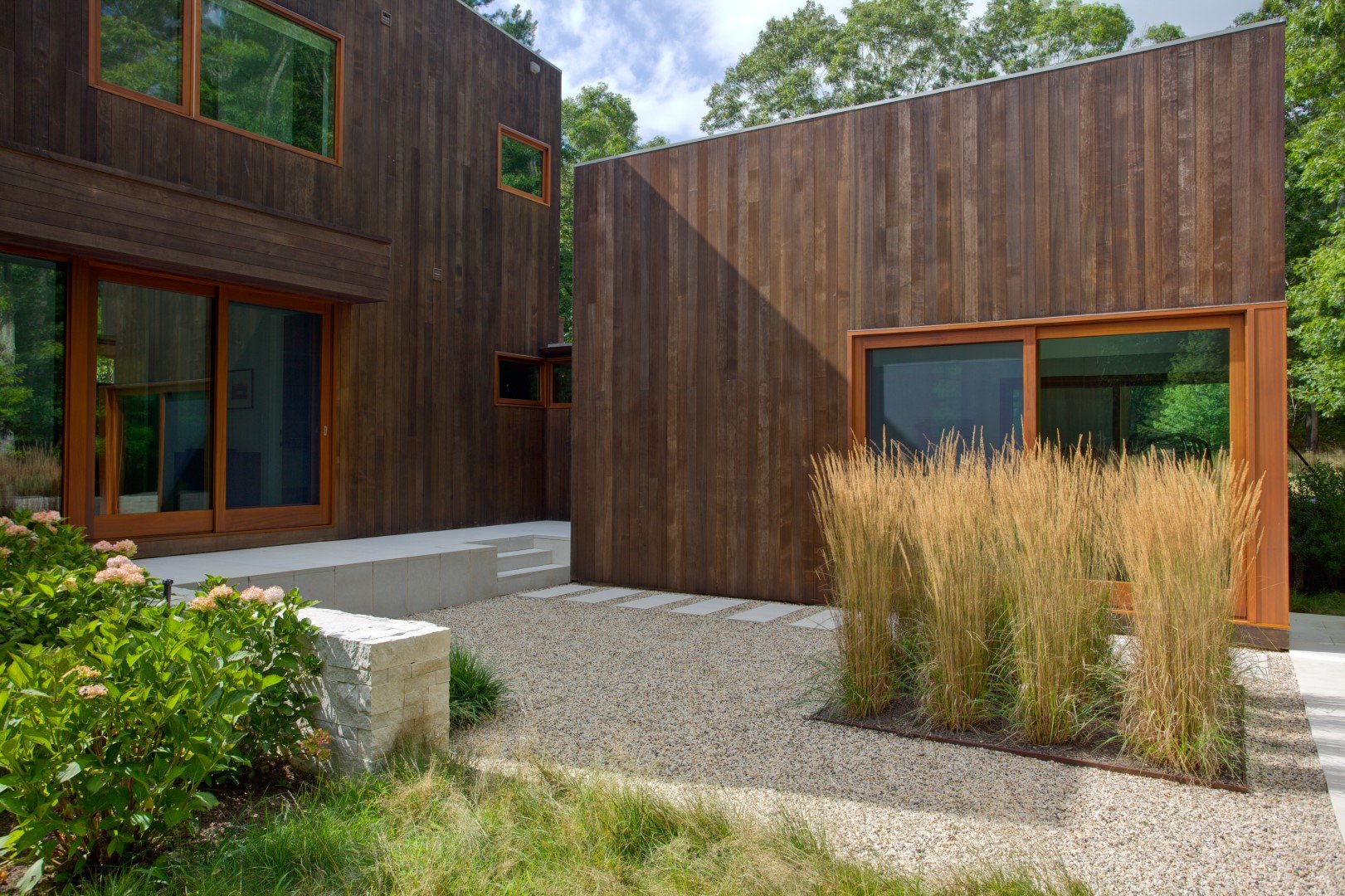 Modern wooden house with large glass windows, surrounded by greenery and tall grass, featuring a gravel walkway and minimal landscaping.