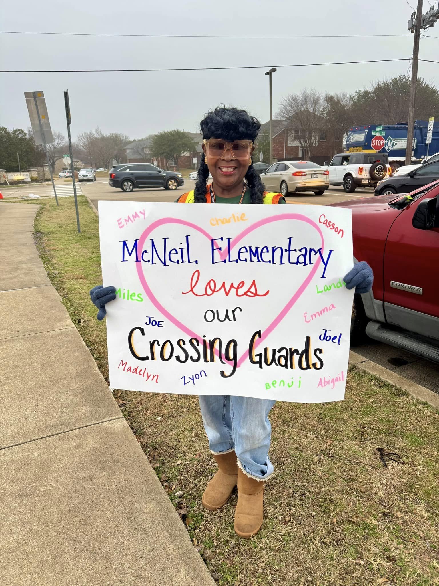 Our crossing guard, who guides students and adults across Hardin Blvd every day, holds a hand made poster board featuring a heart and the message "McNeil Elementary LOVES our Crossing Guards!"