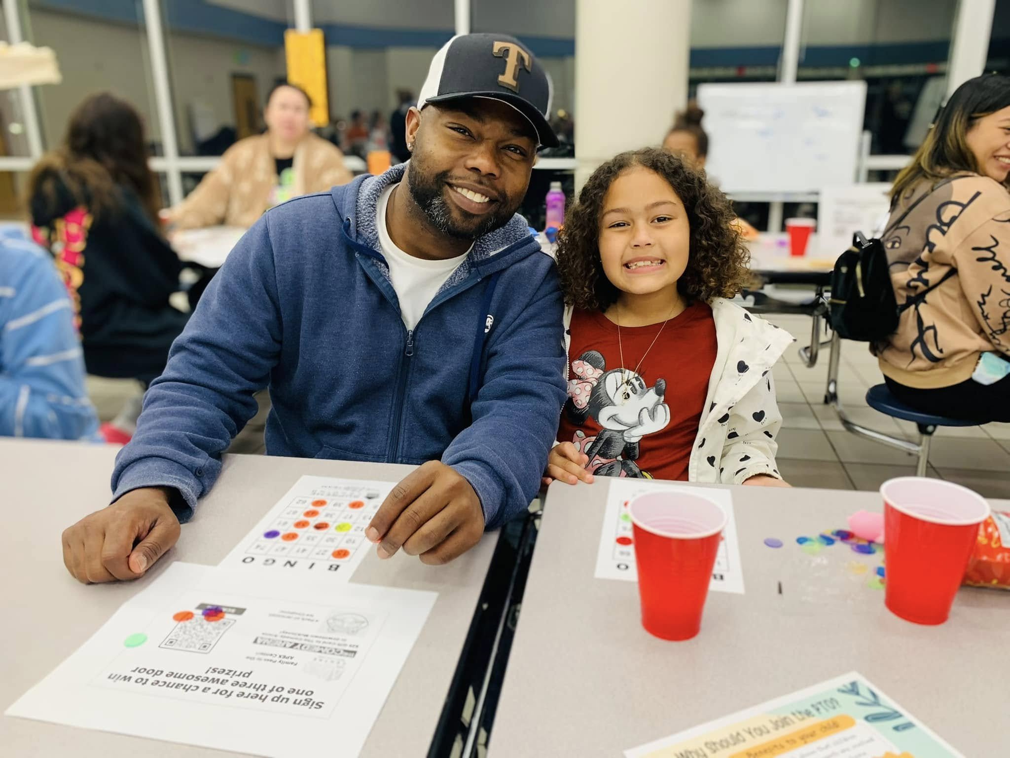 A parent and student seated at a table smile with their Bingo cards.