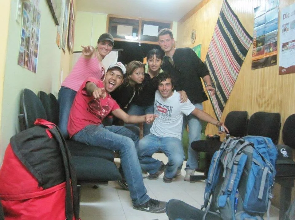 Grupo de seis jóvenes sonriendo y posando en un salón con mochilas y sillas.