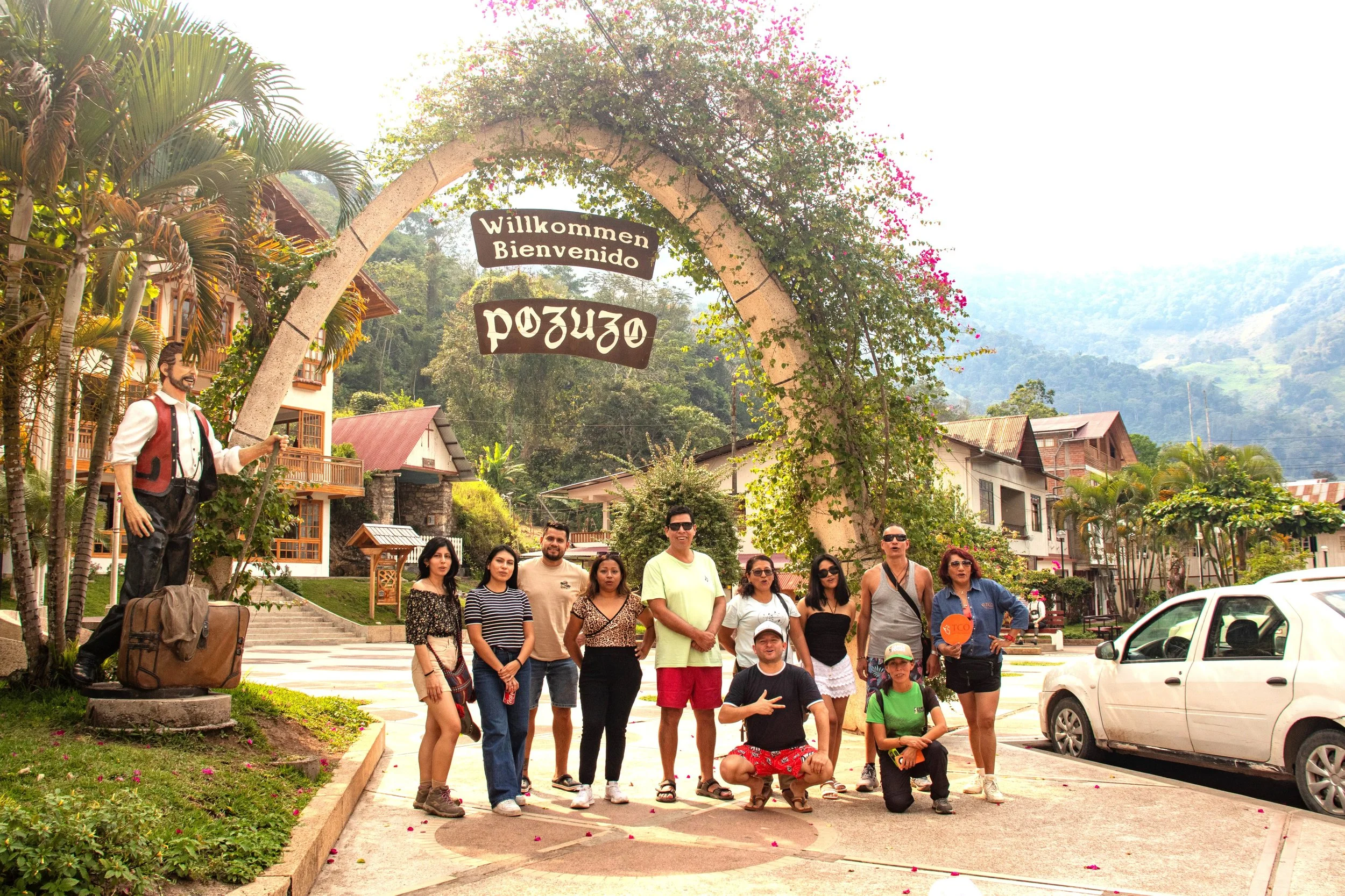 Grupo de turistas posando frente a una señal de bienvenida en un pueblo rodeado de montañas y vegetación, con edificios de madera y carros estacionados.