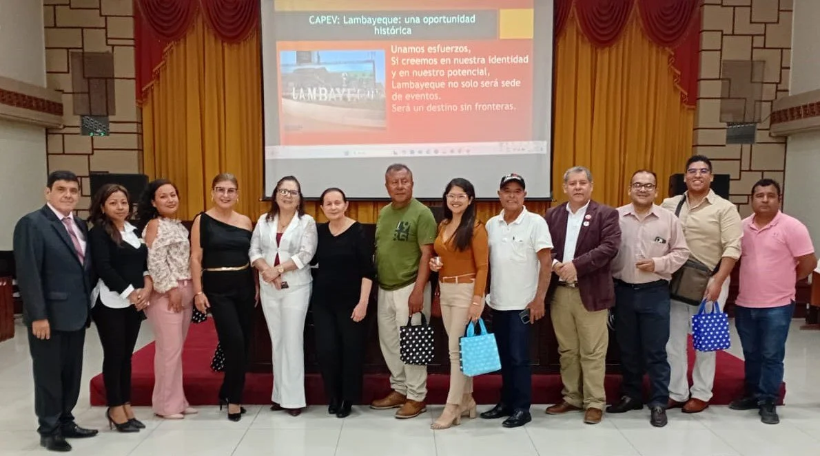 Grupo de personas en un evento, de pie frente a una pantalla con una presentación sobre Lambayeque. Algunos llevan bolsos o bolsas, y todos posan para la fotografía en un salón con cortinas doradas y paredes decoradas.