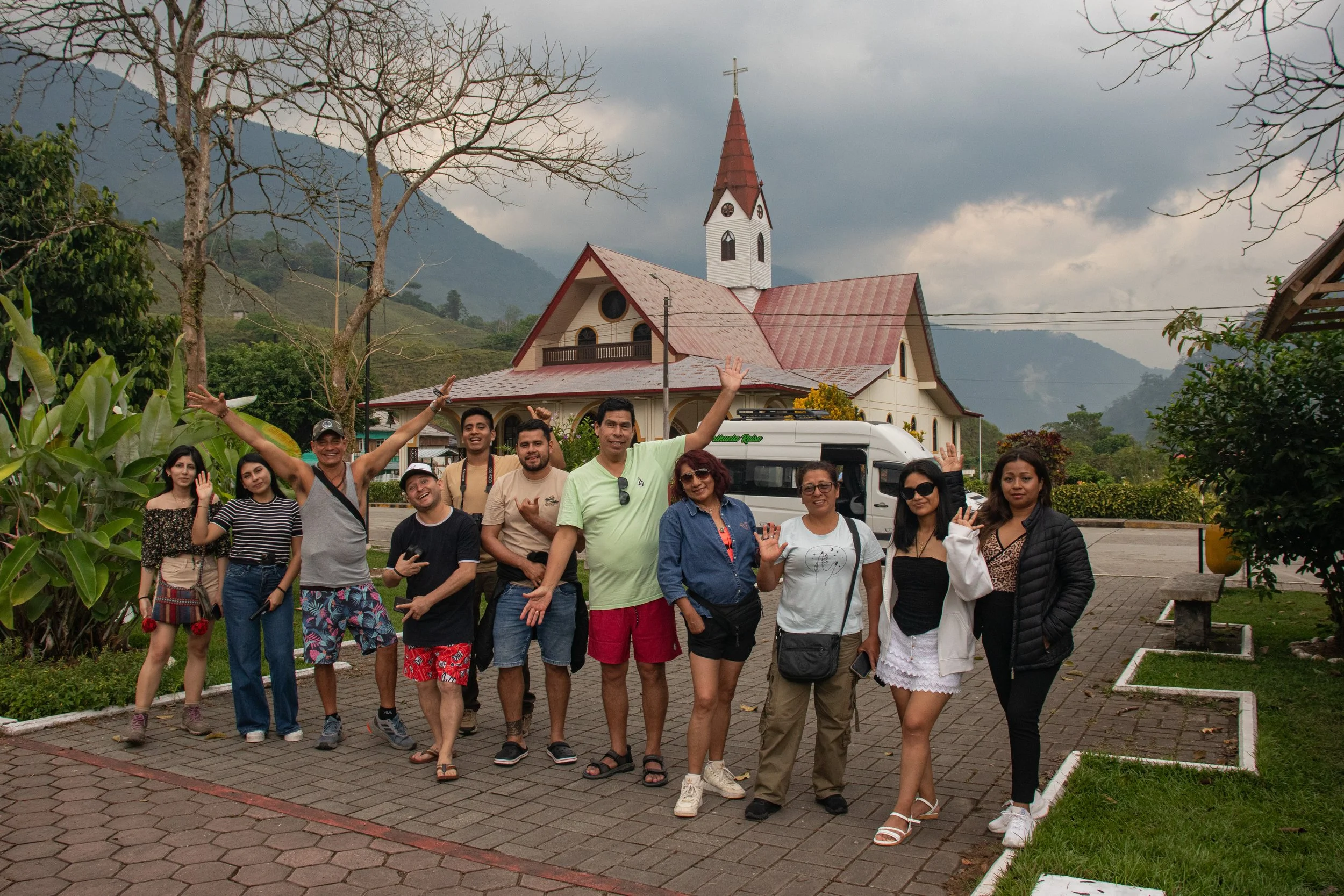 Grupo de personas posando en una calle frente a una iglesia blanca con techo rojo y un cielo nublado, rodeados de árboles y montañas en el fondo.