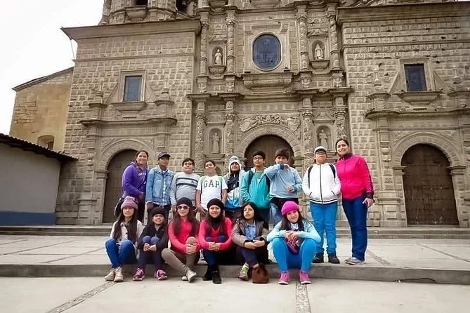Grupo de niños y una adulta posando frente a una iglesia antigua de piedra en una plaza.