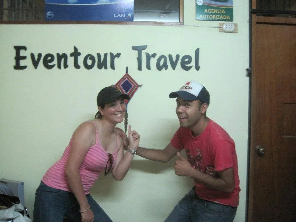 Dos personas sonriendo y señalándose, en una oficina con cartel que dice 'Eventour Travel' y un letrero adicional que indica 'Agencia Autorizada'.