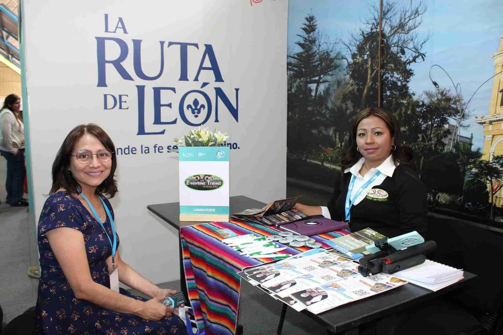 Dos mujeres en un stand en una feria, una de ellas con gafas y vestido azul con flores, y otra con blusa blanca y chaqueta negra, en un fondo con un árbol y edificios, junto a una mesa con folletos, papelería y objetos promocionales.