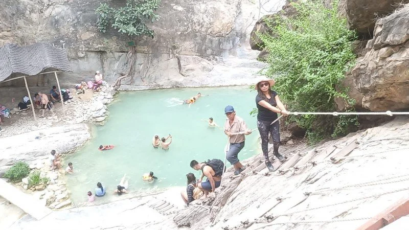 Personas en una piscina natural rodeada de rocas y vegetación, algunas personas nadando y otras en el borde de la piscina.
