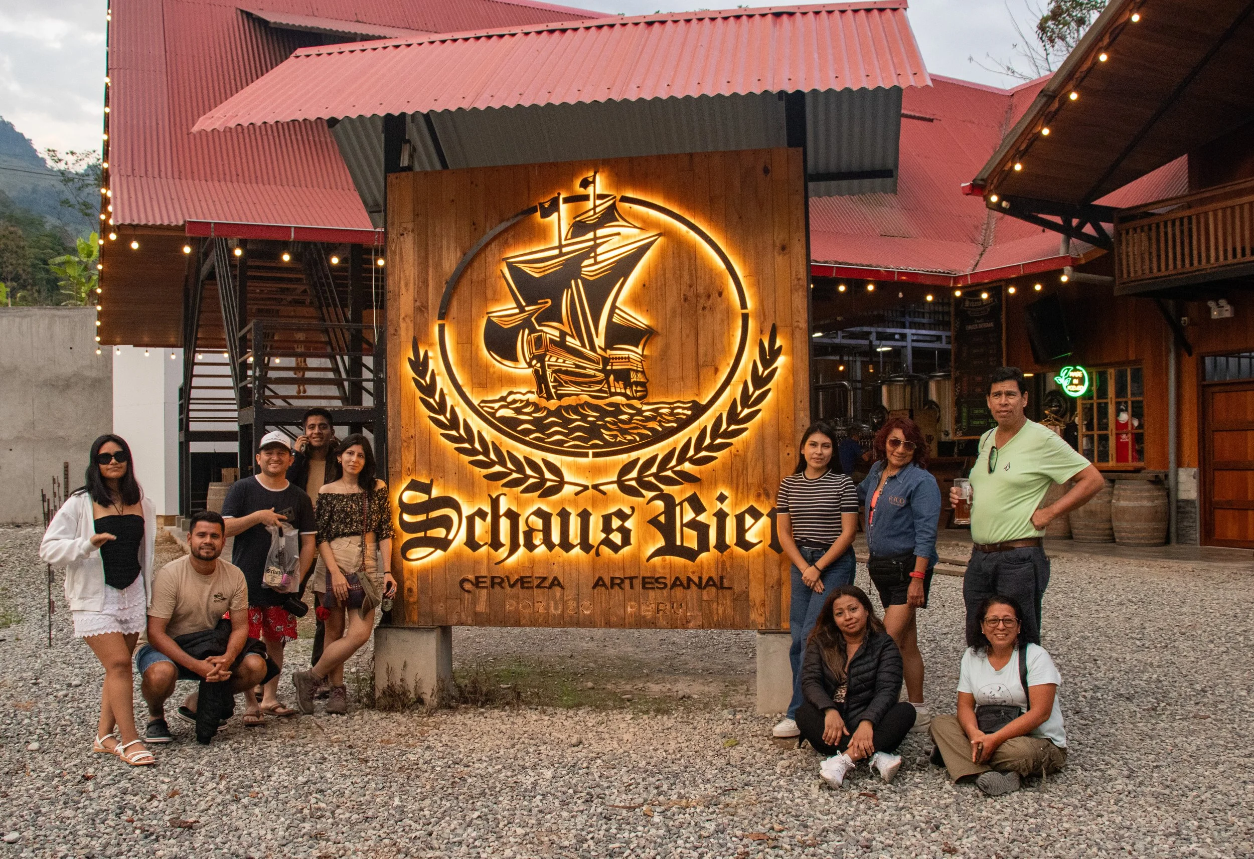 Grupo de personas posando frente a un cartel de madera con el logotipo de shirkook, que representa un barco pirata, y el texto "Sharks Bier" y "Cerveza Artesanal" en un establecimiento con paredes de madera y techo de lámina, decorado con luces y plantas.