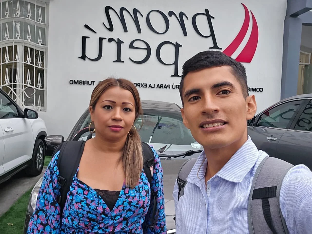 Dos personas, un hombre y una mujer, toman una selfie frente a un edificio con cartel que dice 'Dora Pérez', en un área con autos estacionados.