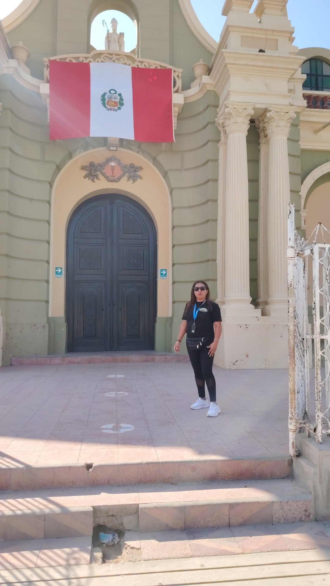 Persona joven de cabello largo y gafas de sol, vestida con camiseta negra y pantalones deportivos negros, posa frente a una fachada de estilo clásico con columnas y un escudo en la parte superior, en un día soleado.