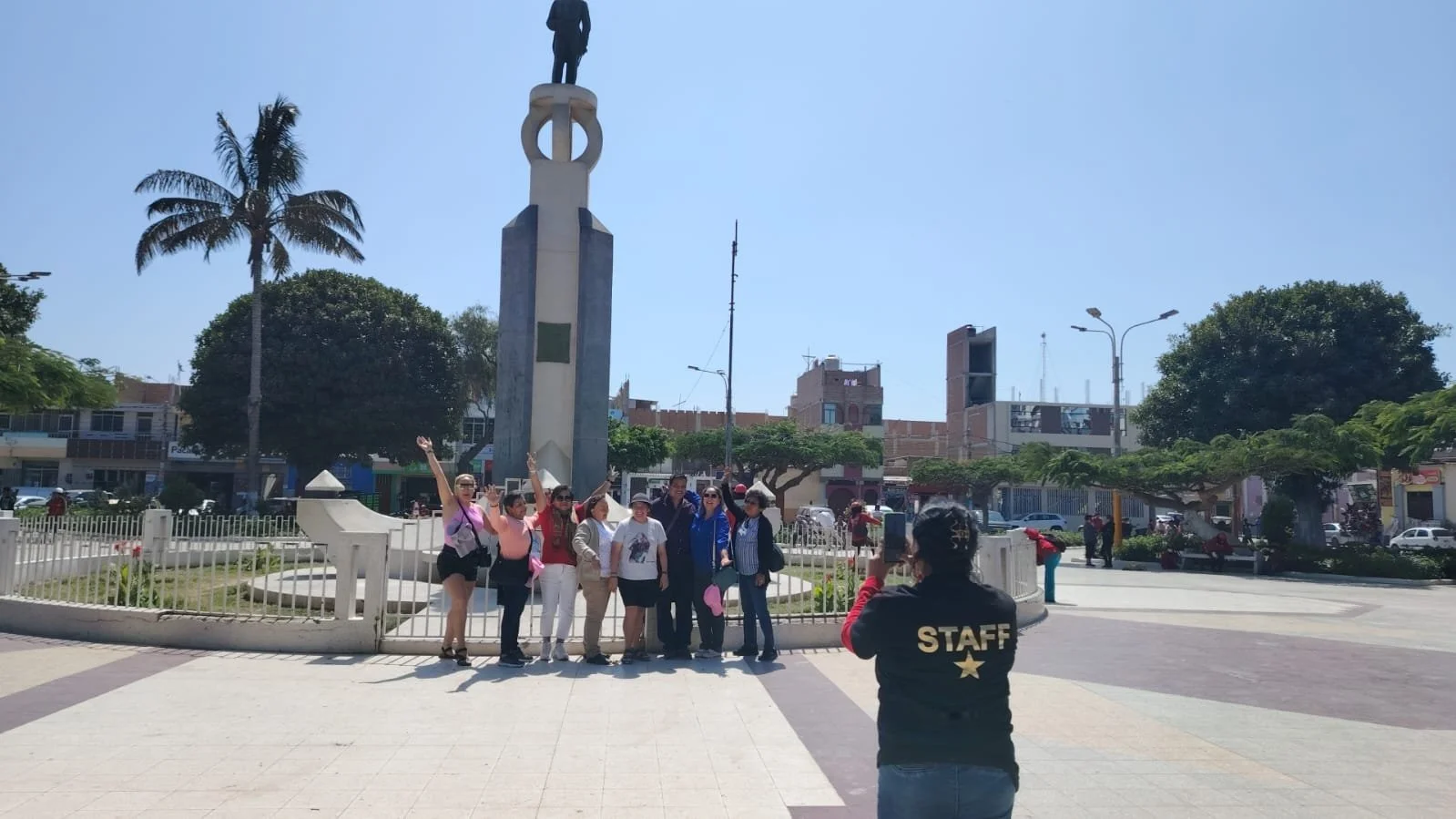 Grupo de personas posando frente a una estatua en una plaza central, una de ellas lleva una camiseta que dice 'STAFF', y otra está tomando fotos.