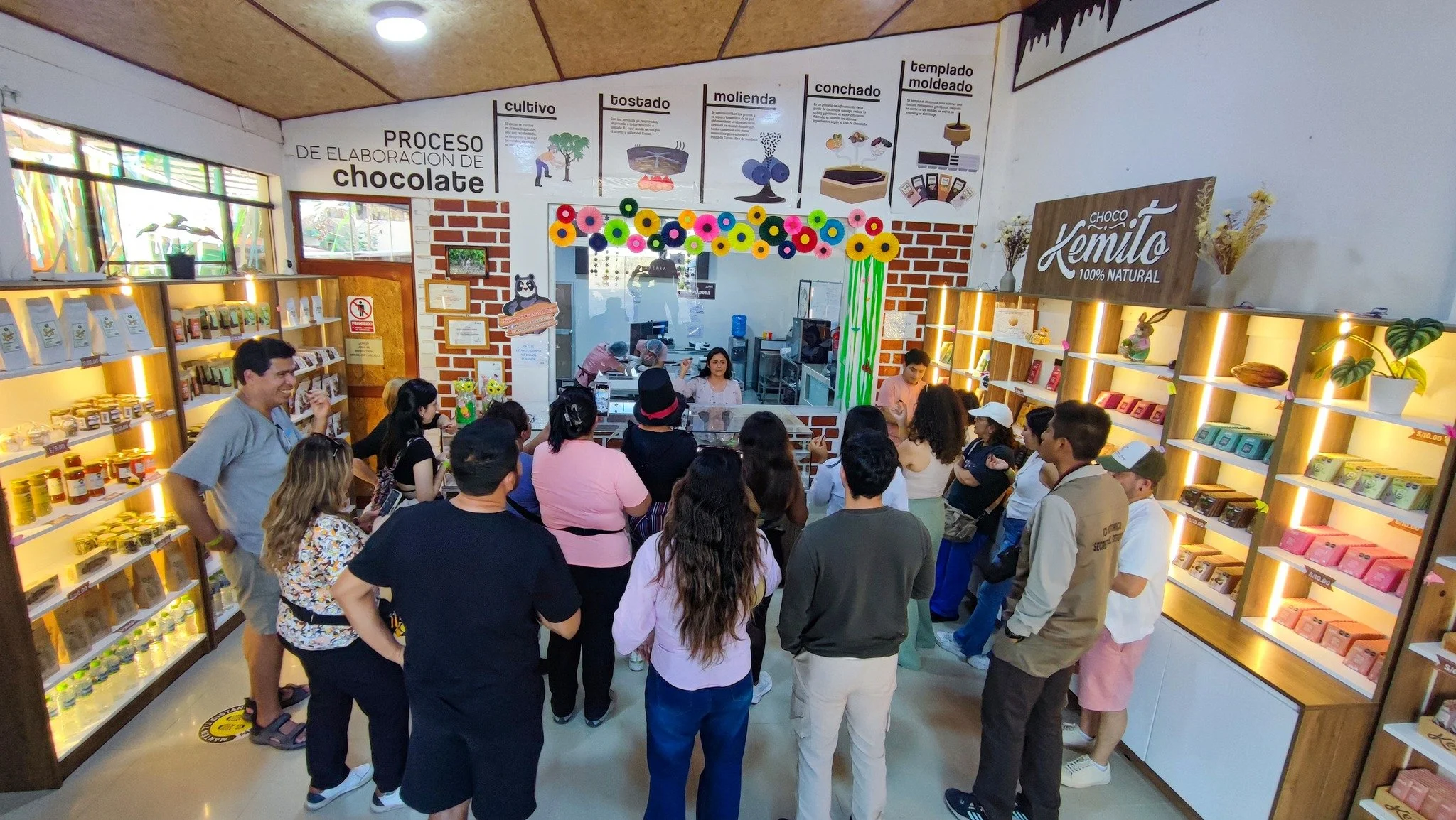 Personas dentro de una tienda de chocolates, con estantes llenos de productos y una pared con información del proceso de elaboración del chocolate.