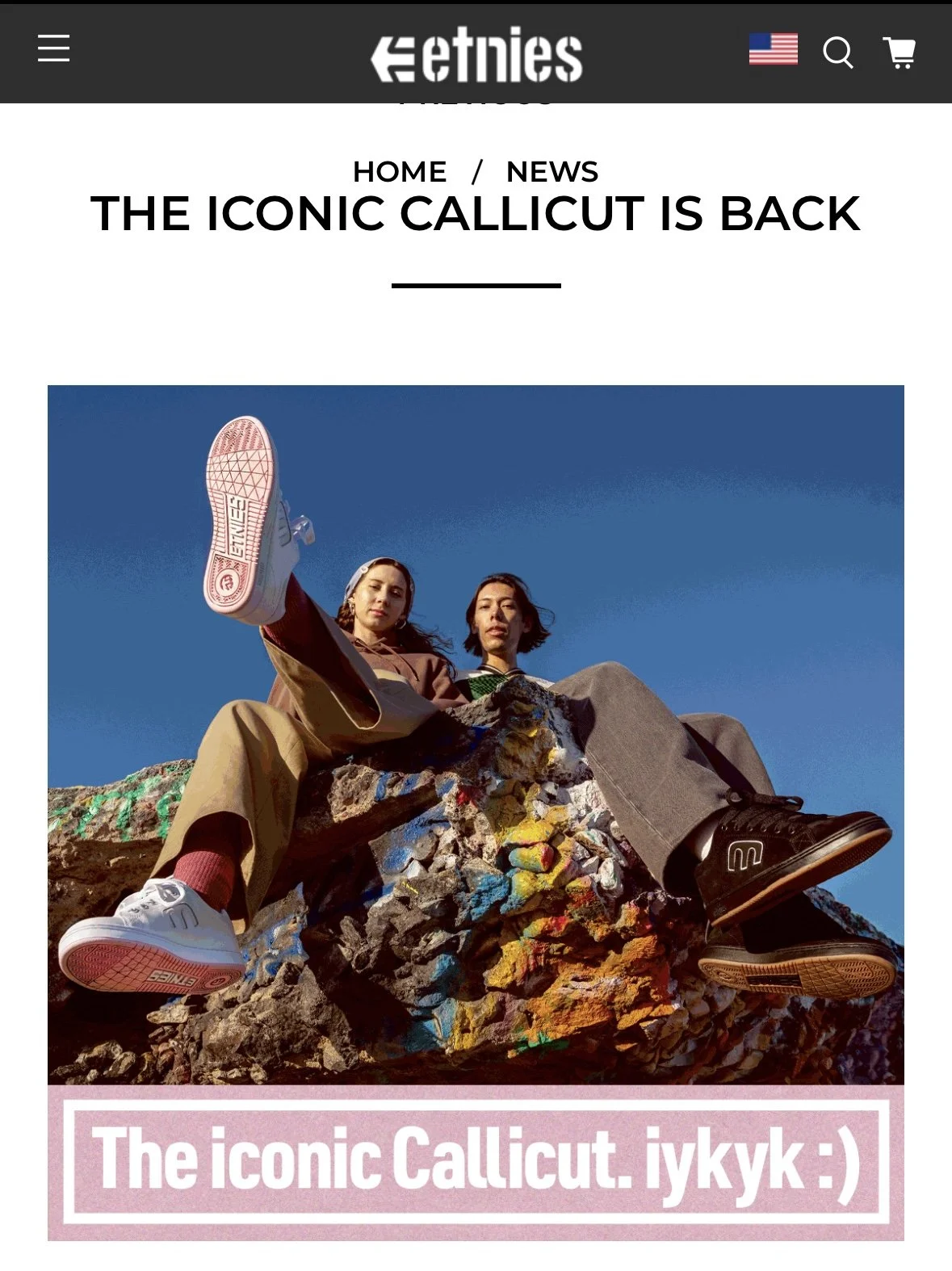 Two women sitting on a colorful rock formation wearing casual clothing and sneakers, with a clear blue sky in the background.