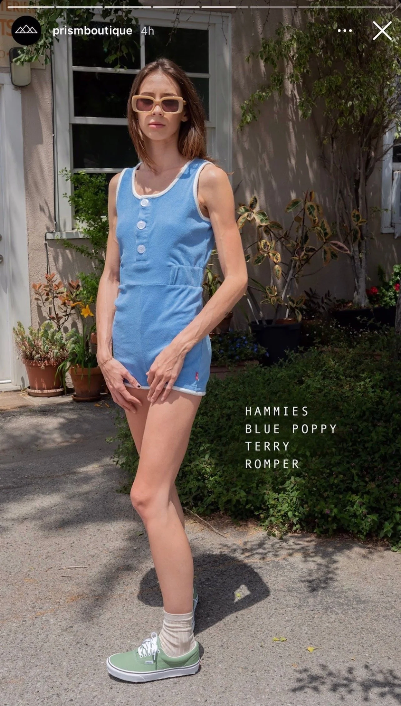 Young woman standing outdoors in front of a house, wearing a blue terry romper, beige socks, and green sneakers with white laces. She has brown hair, sunglasses, and is posing with her hands near her hips. Potted plants and a window are visible in the background.