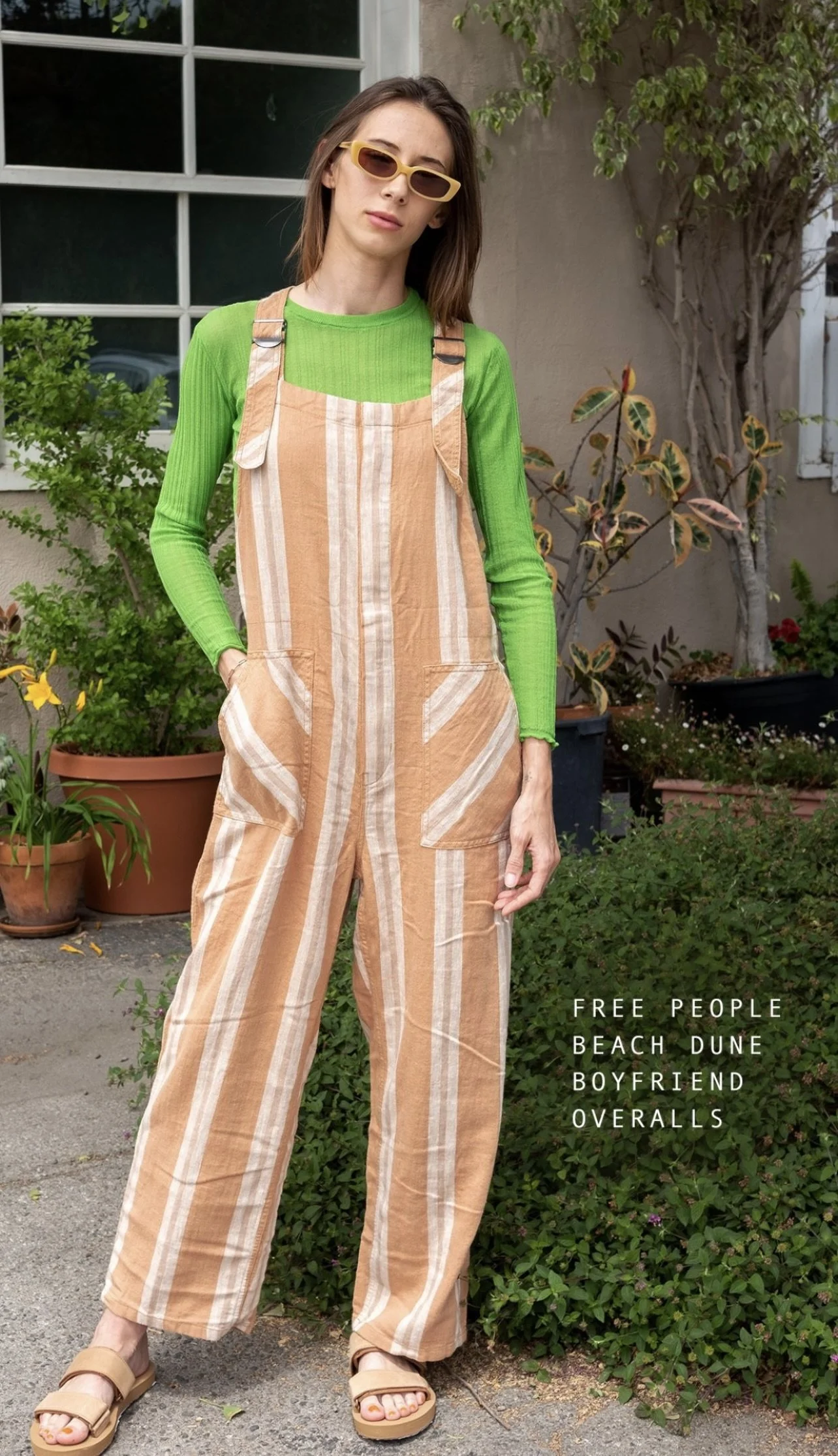 A young woman with shoulder-length brown hair wearing sunglasses, a bright green long-sleeve shirt, and beige striped overalls, standing outdoors in a garden with potted plants and a gray wall.