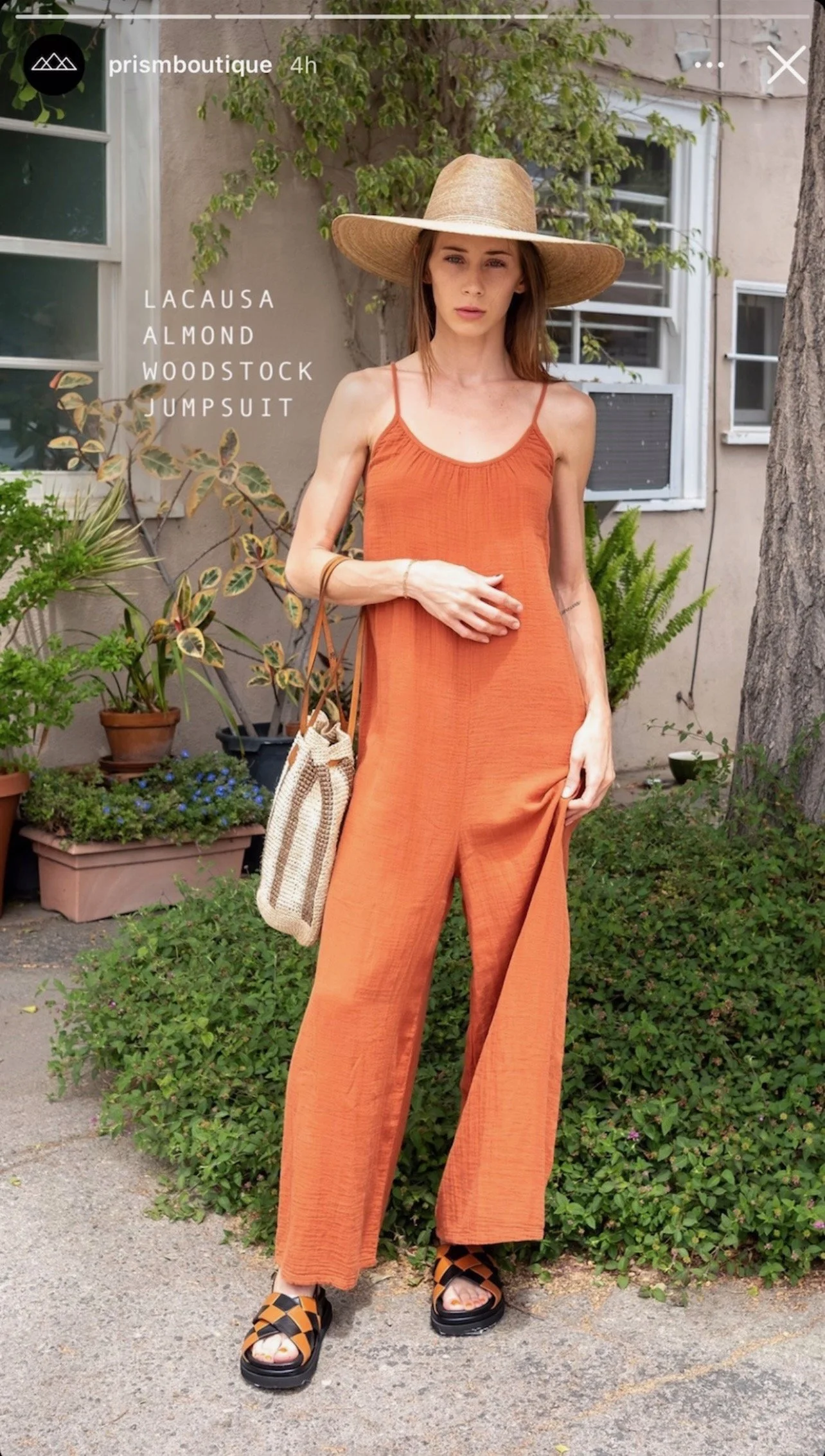 A woman wearing an orange Lacausa almond-colored Woodstock jumpsuit, a wide-brimmed straw hat, black and orange sandals, and holding a beige woven tote bag, standing outdoors next to greenery and a building.
