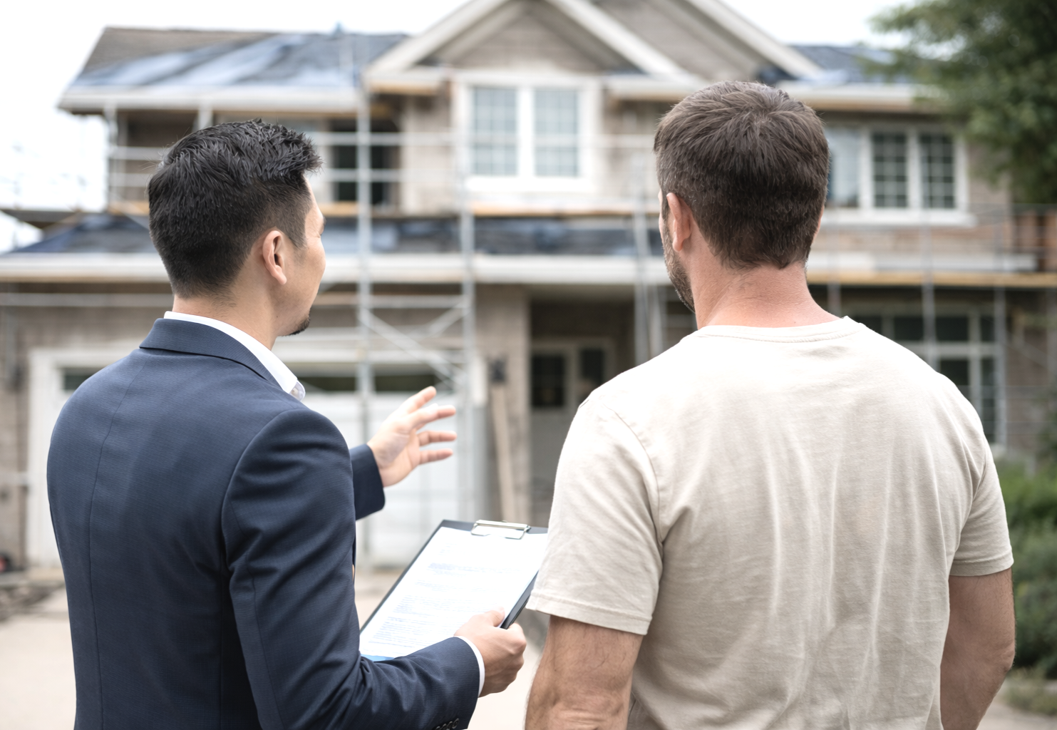 Two men, one in a suit and one in a casual t-shirt, standing in front of a house under construction. The man in a suit appears to be explaining something, holding a clipboard.