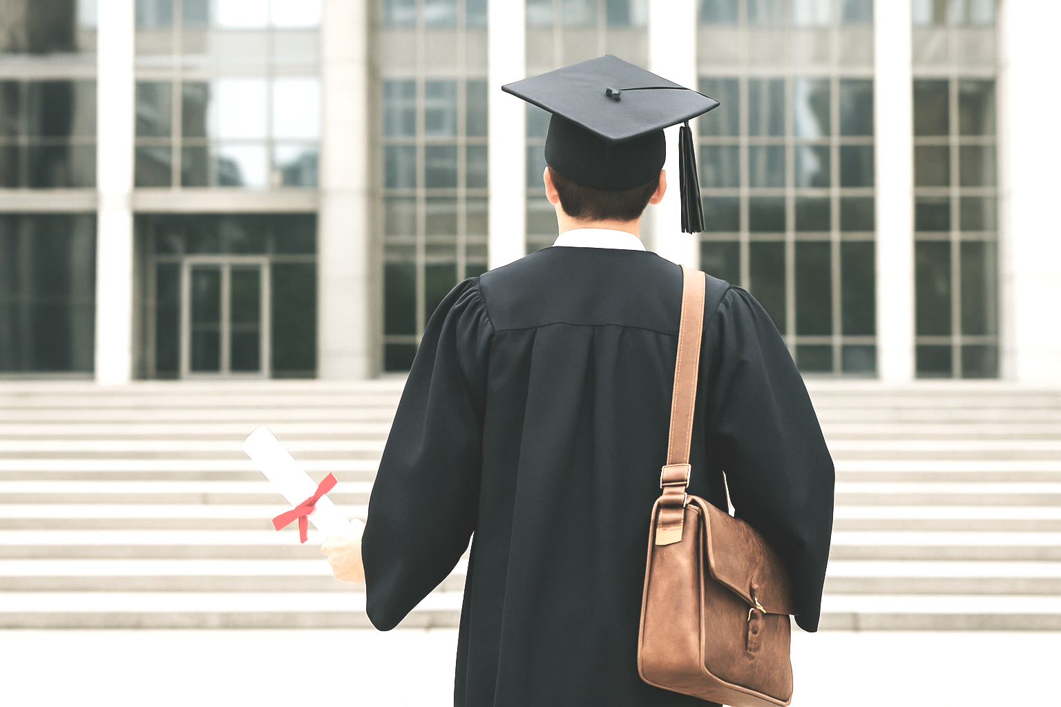 A person dressed in a black graduation gown and cap holding a diploma, walking toward a modern building with large glass windows and steps.