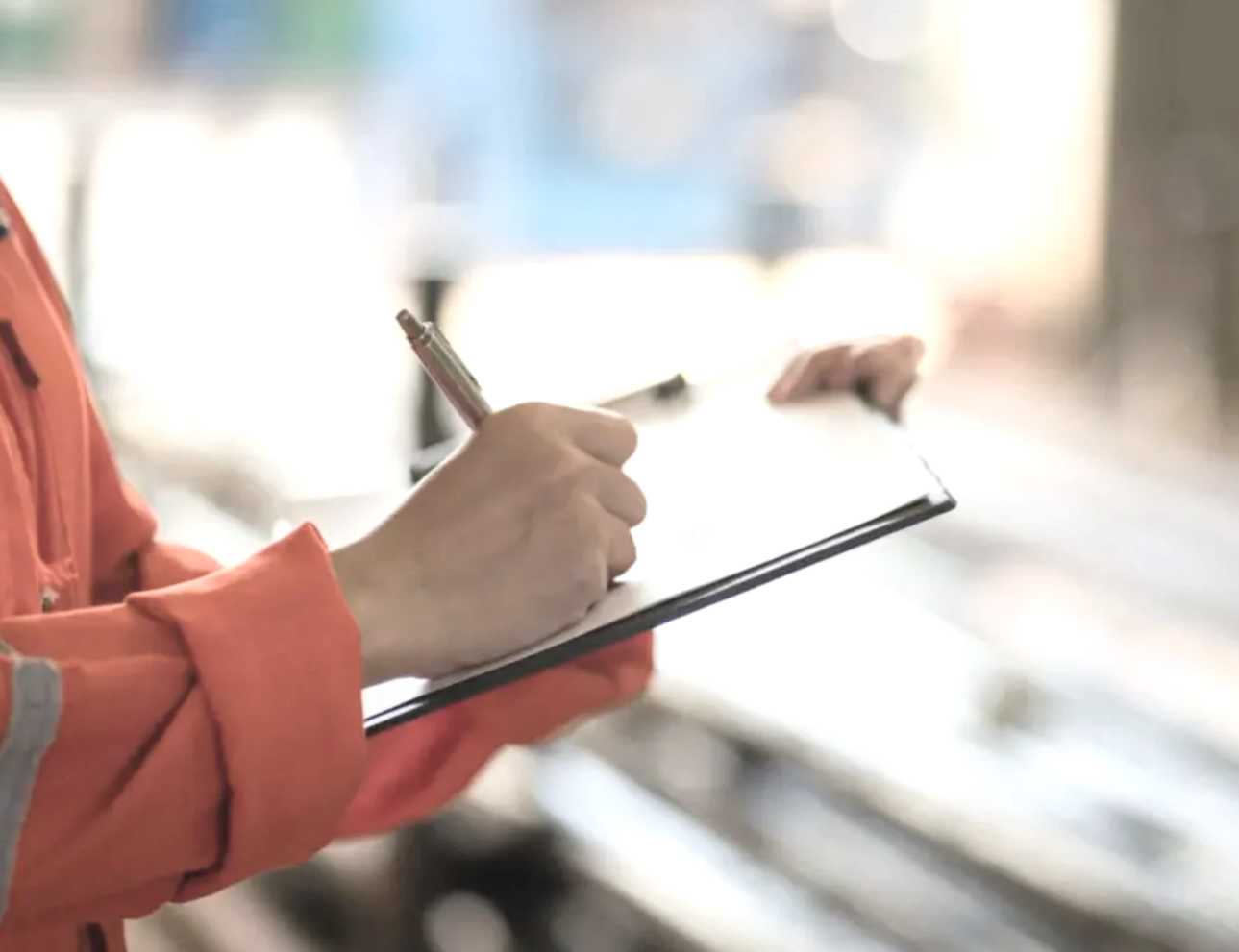 Person in an orange jacket writing on a notepad with a pen.
