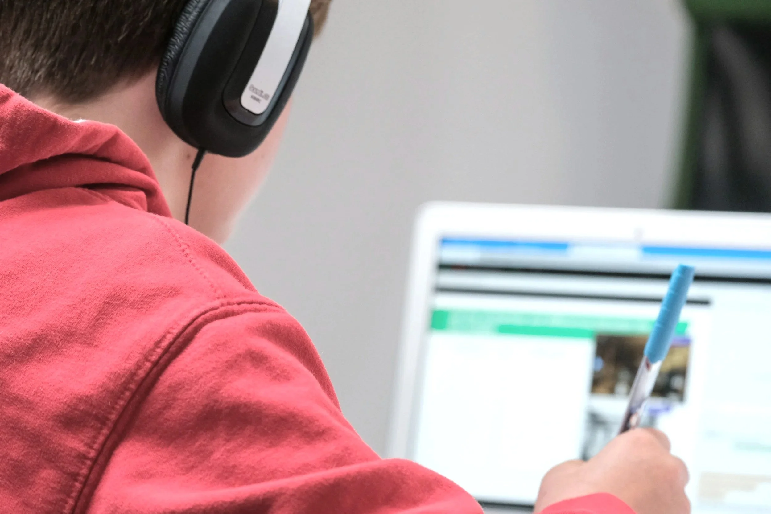 A person wearing a red hoodie and headphones is sitting at a desk, using a laptop and holding a pen, with a spreadsheet or document visible on the screen.
