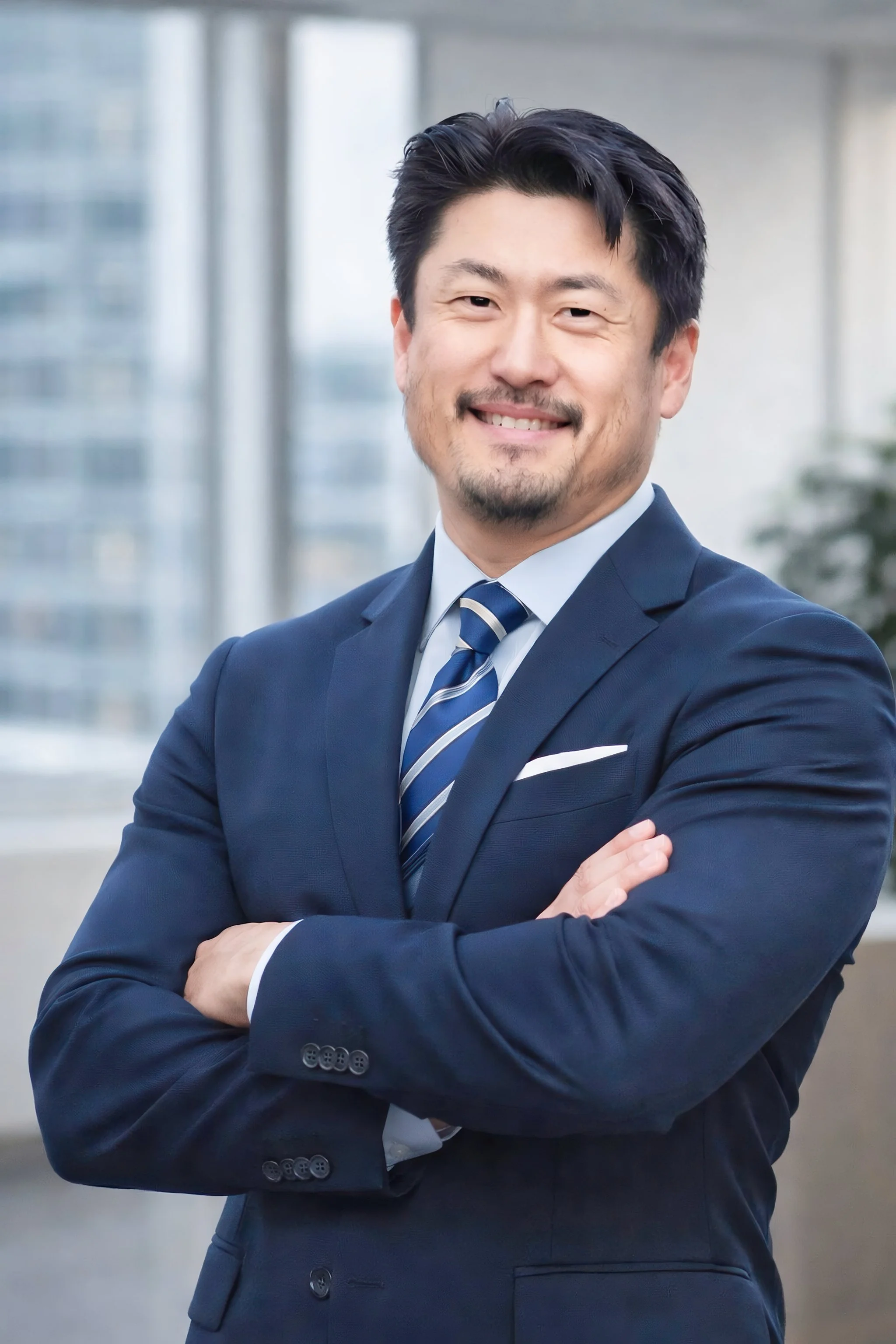 A confident Asian man smiling with arms crossed, dressed in a navy suit, white shirt, and striped tie, standing in front of a modern office building.