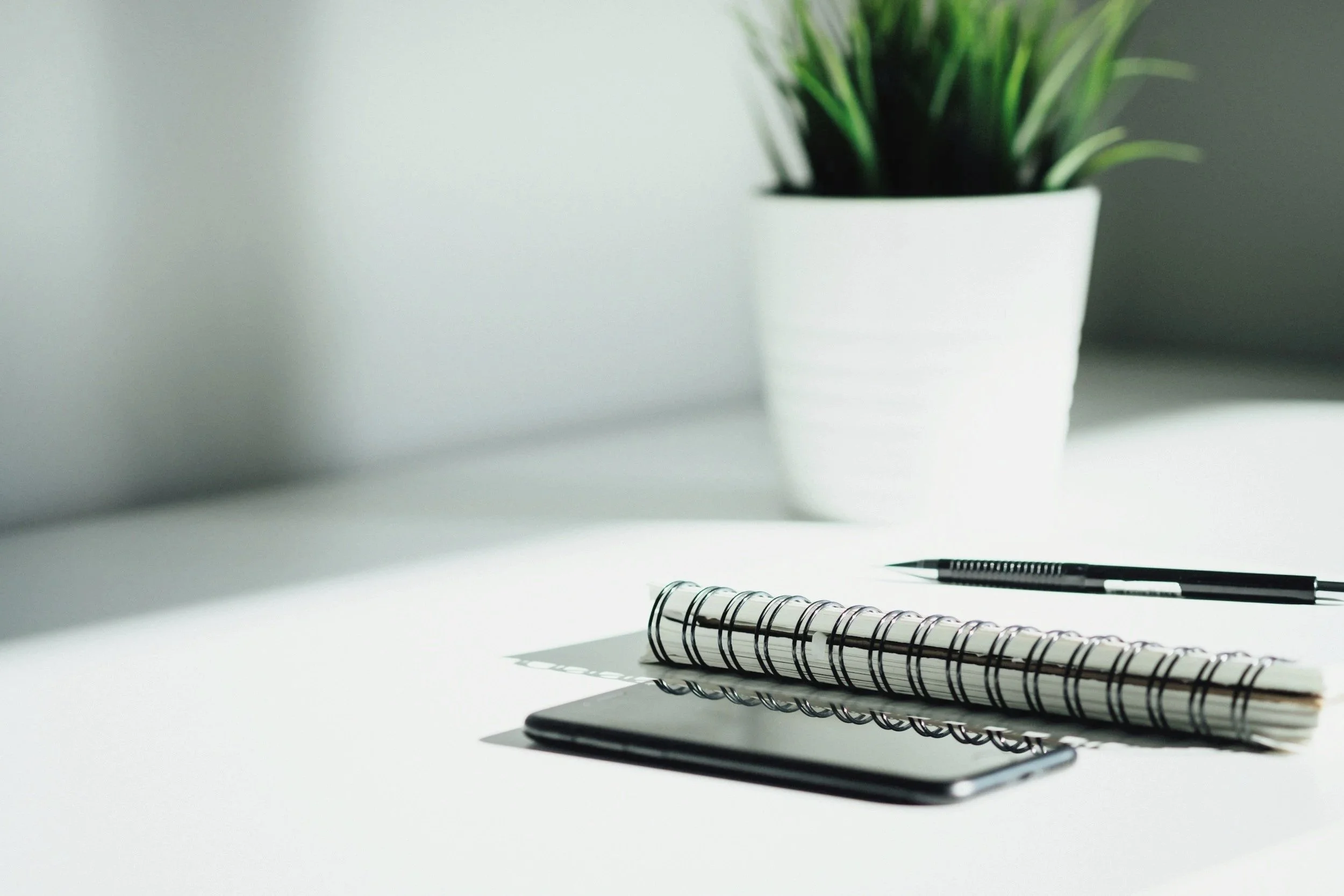 A minimalist desk setup with a potted green plant, a black pen, a closed tablet, a spiral notebook, and a smaller notebook on a white surface with soft lighting.