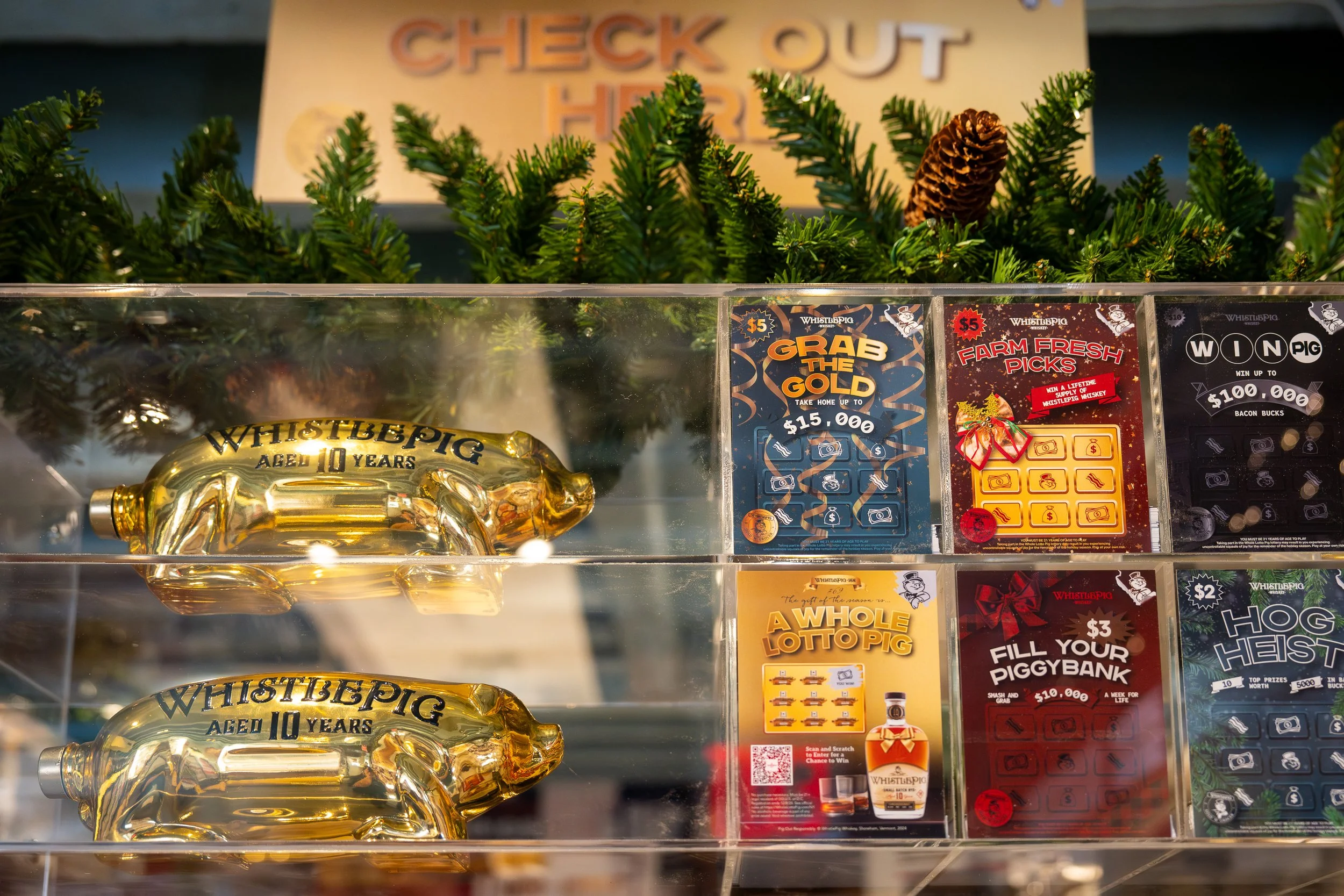 Display case with two gold pig-shaped bottles labeled 'Whistlepig Aged 10 Years' and promotional signs for lottery-style tickets and a bottle of Whistlepig whiskey, surrounded by Christmas greenery.