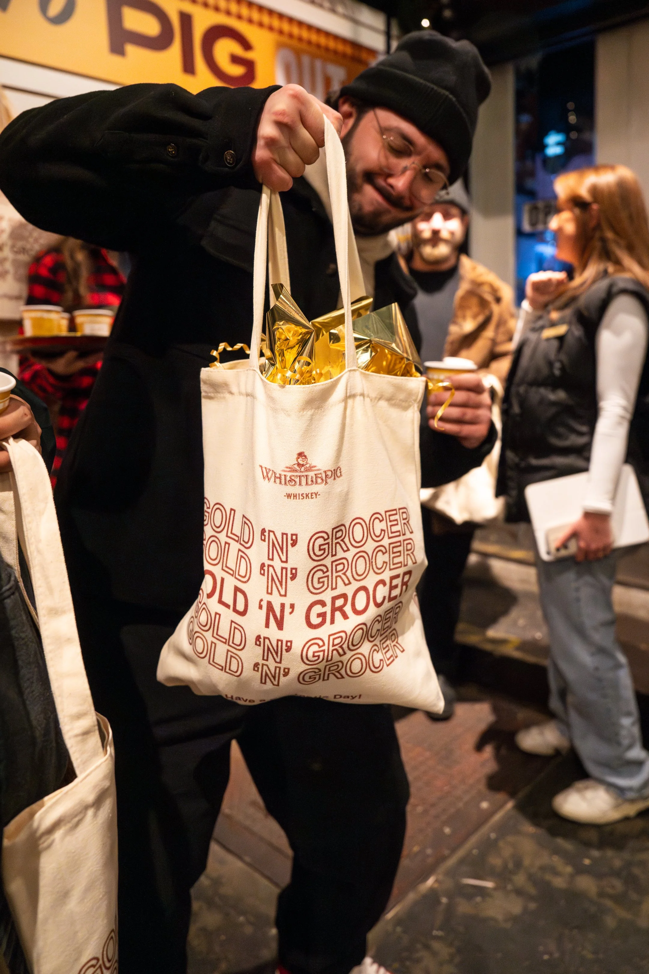 A man in a black coat and beanie holding a tote bag filled with gold-wrapped gifts and candy. People stand in the background outside a shop with a sign that reads "BIG".