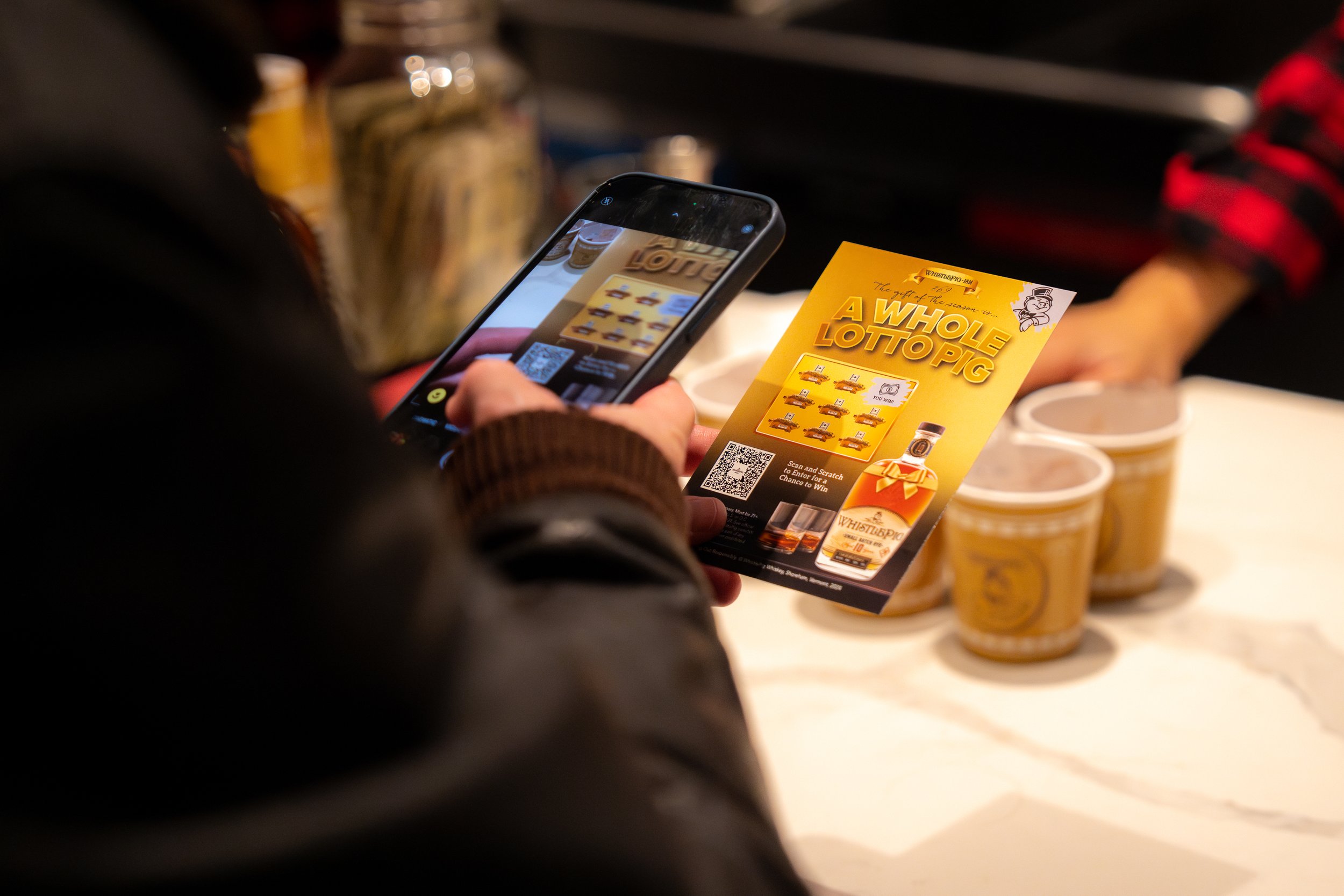 Person taking a photo of a yellow promotional flyer titled 'A WHOLE LOTTO PIG' with their smartphone. The flyer features images of whiskey bottles and a QR code. There are three whiskey bottles on the table in the background.