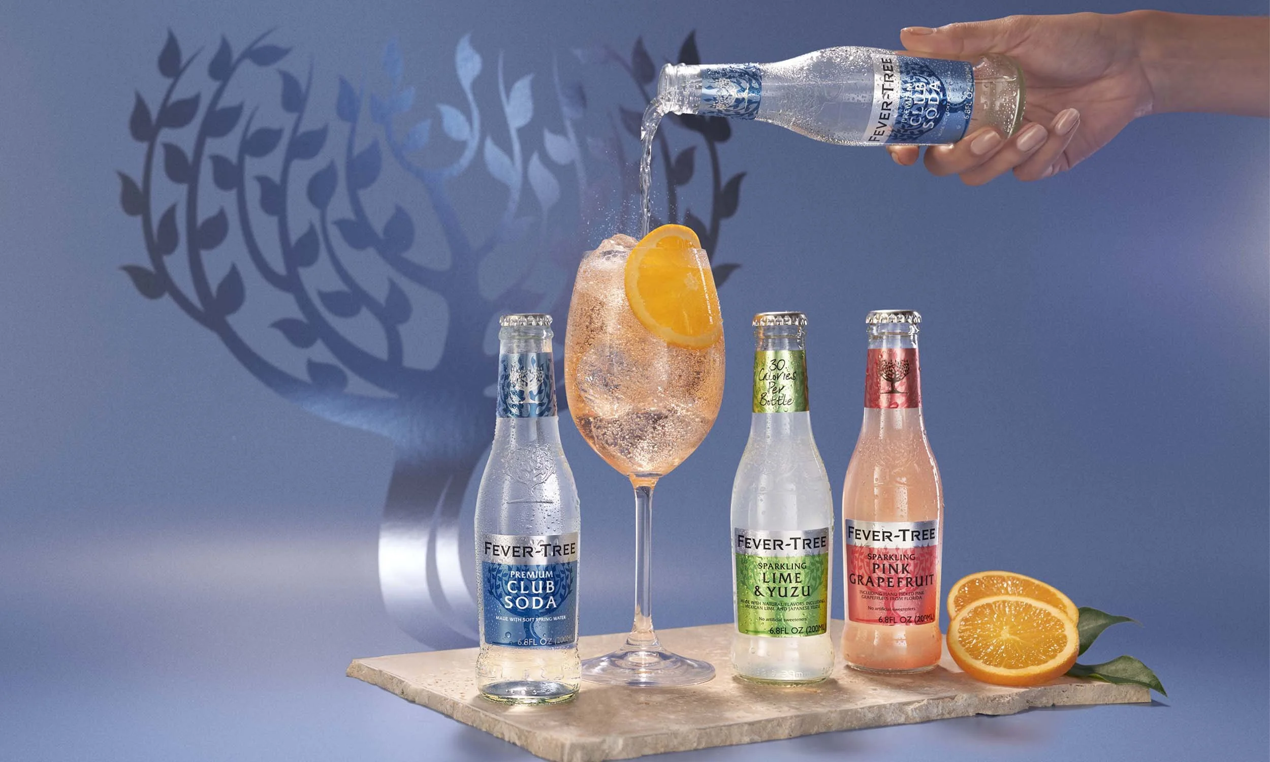 A hand pouring clear sparkling water into a wine glass filled with pink soda and ice, garnished with an orange slice, on a wooden surface with three bottles of soda and a halved orange with leaves, against a blue background with a shadow of a tree.