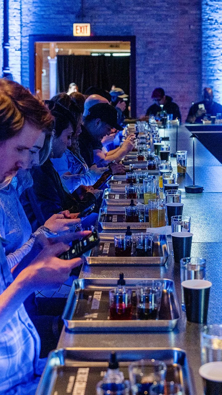 People sitting at a bar tasting different colorful liquids with equipment, in a dimly lit, modern space.