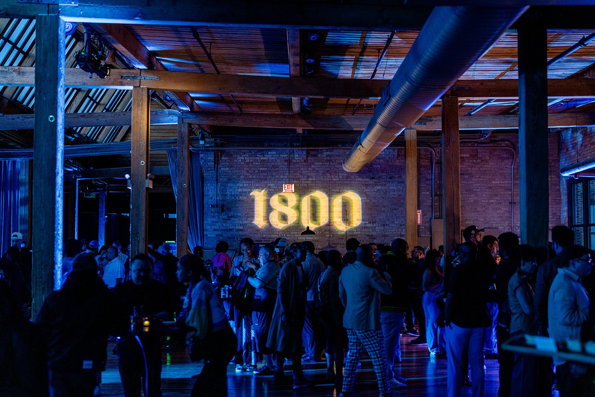 Indoor event with a crowd of people socializing, illuminated by blue lighting, with the number 1800 projected on a brick wall.