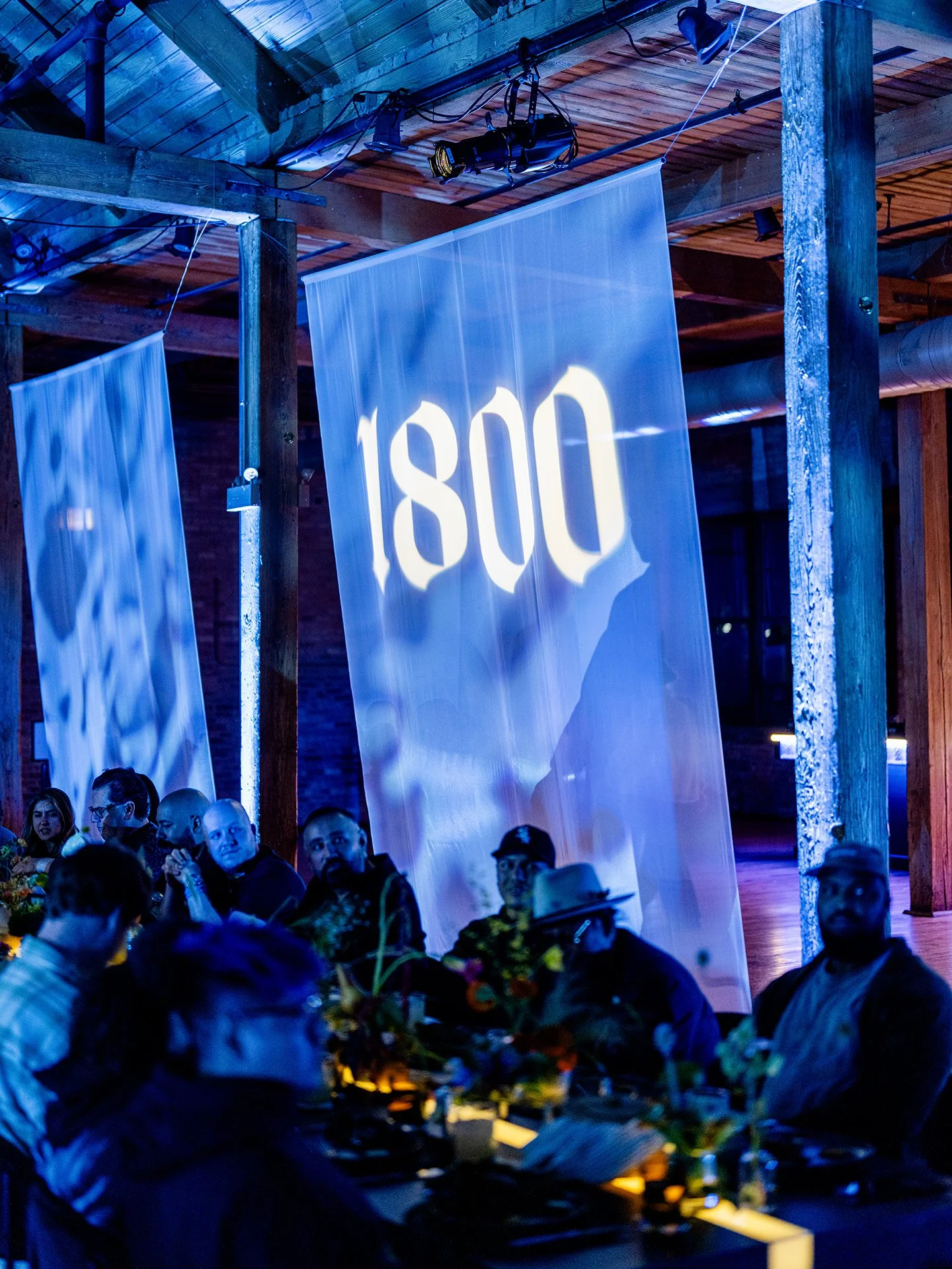 People sitting at a decorated banquet table in a dimly lit venue with large banners displaying 1800. The scene has a wooden ceiling and walls, with stage lighting creating a vibrant atmosphere.