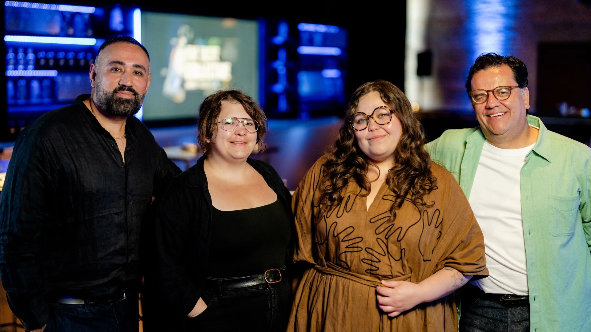 Four taste experts, including Dr. Arielle Johnson, Chef Ana Castro, bartender Nacho Jimenez, and agave expert Jaime Salas, are standing together in a dimly lit room with a bar and TV screen in the background, smiling at the camera.